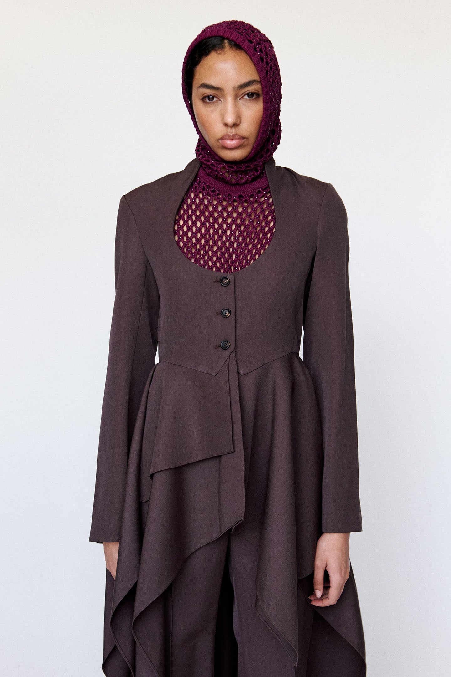 Model is wearing a dark brown jacket with a open chest and 3 button closure paired with a crystal mesh magenta top and crystal mesh magenta hood with a flowy layered brown skirt and matching brown pants photographed against a white background