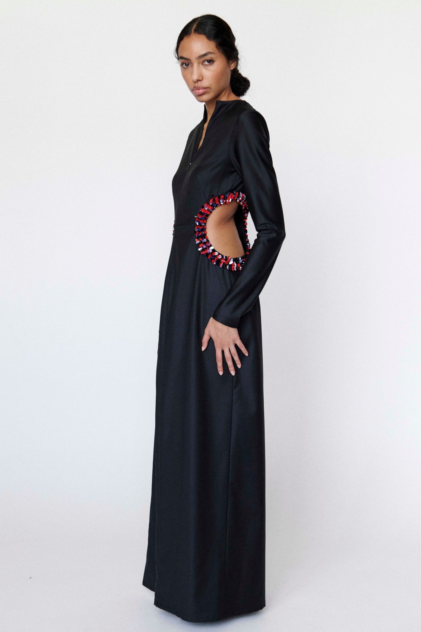Model posing to the side is wearing a black dress with colorful circular cutout design. the cutout design is trimmed with a red, black, and silver palettes design on a white background 