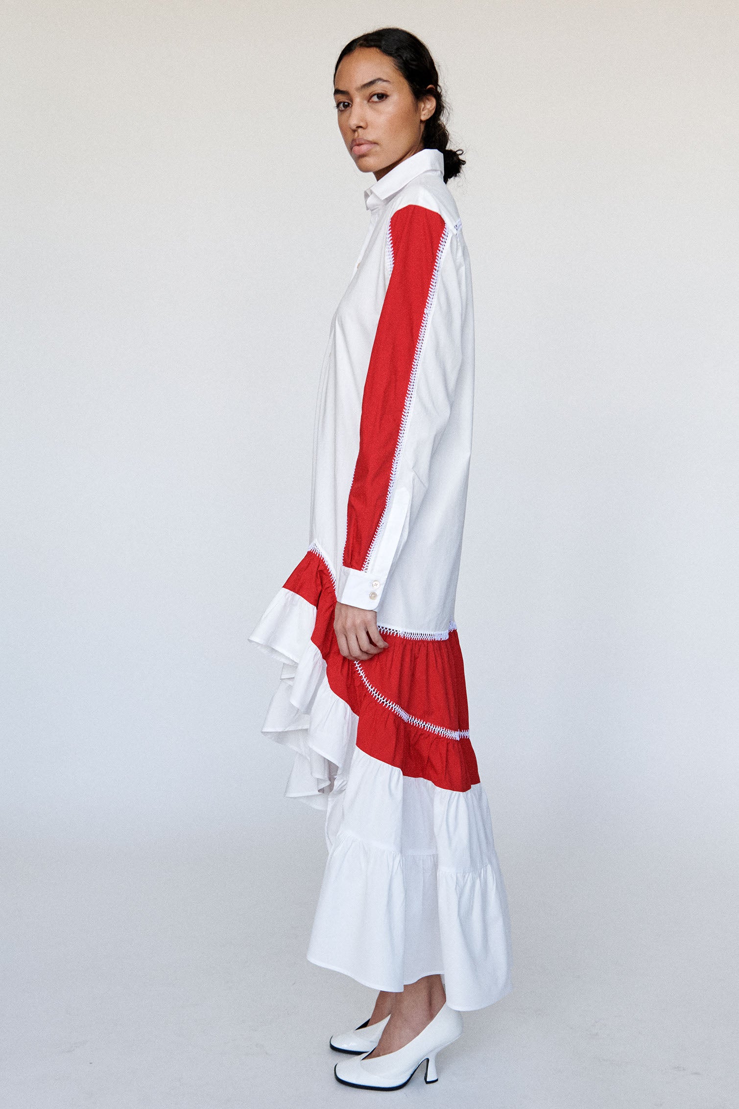 Model wears white skirt dress with hand stitched red accent panelling and gathered ruffle hem with white pump. 