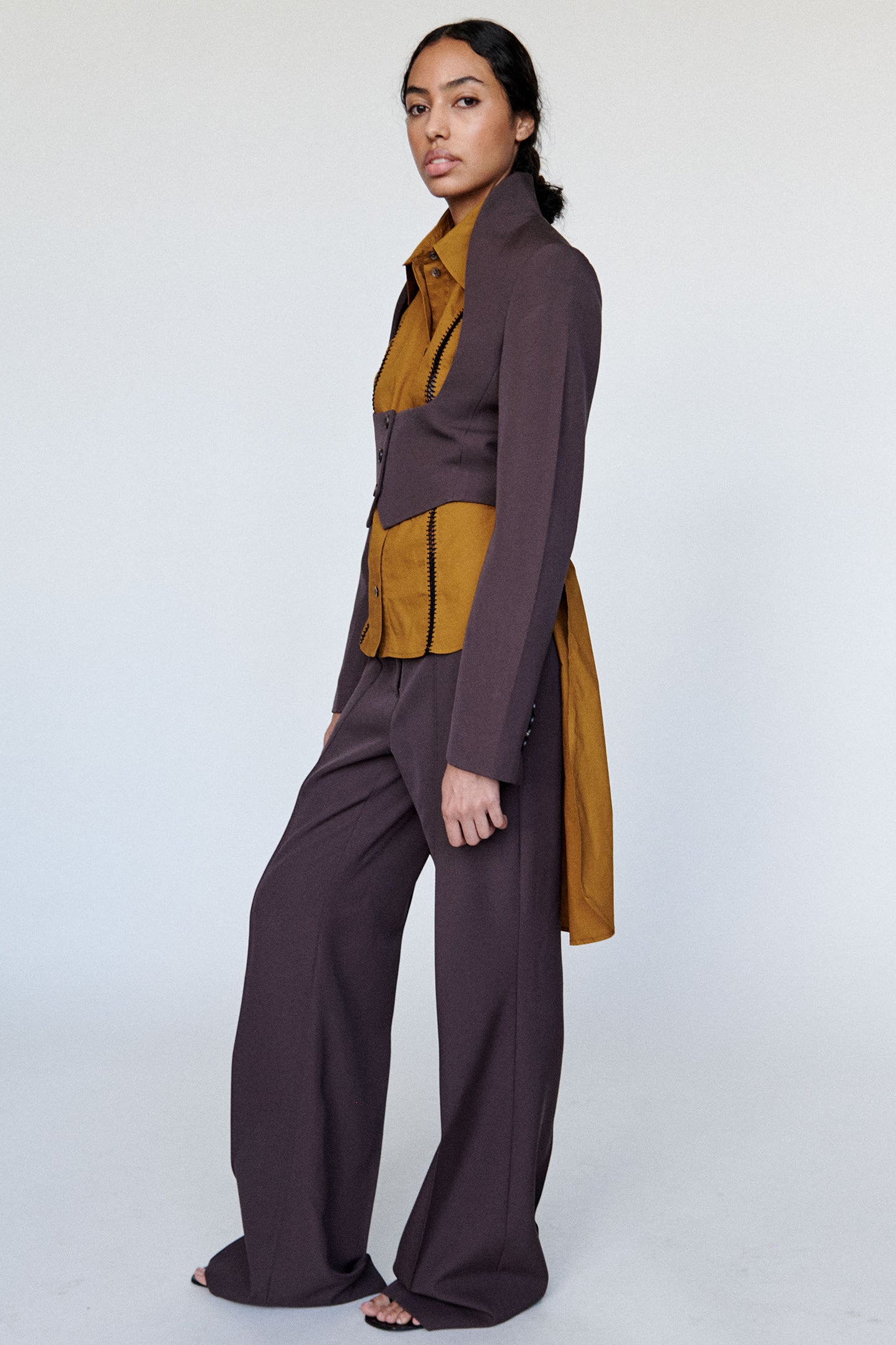 Model is wearing a brown relaxed pants with a mustard button down top with a fitted brown open chest jacket over with a three button closure photographed on a white background