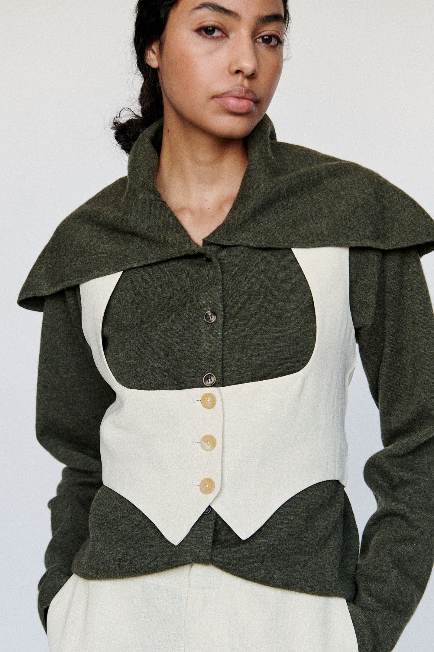 Model wears green-grey cashmere cardigan with exaggerated sailor collar. An off-white vest is worn on top on the cardigan with the sailor collar pulled over the vest. 