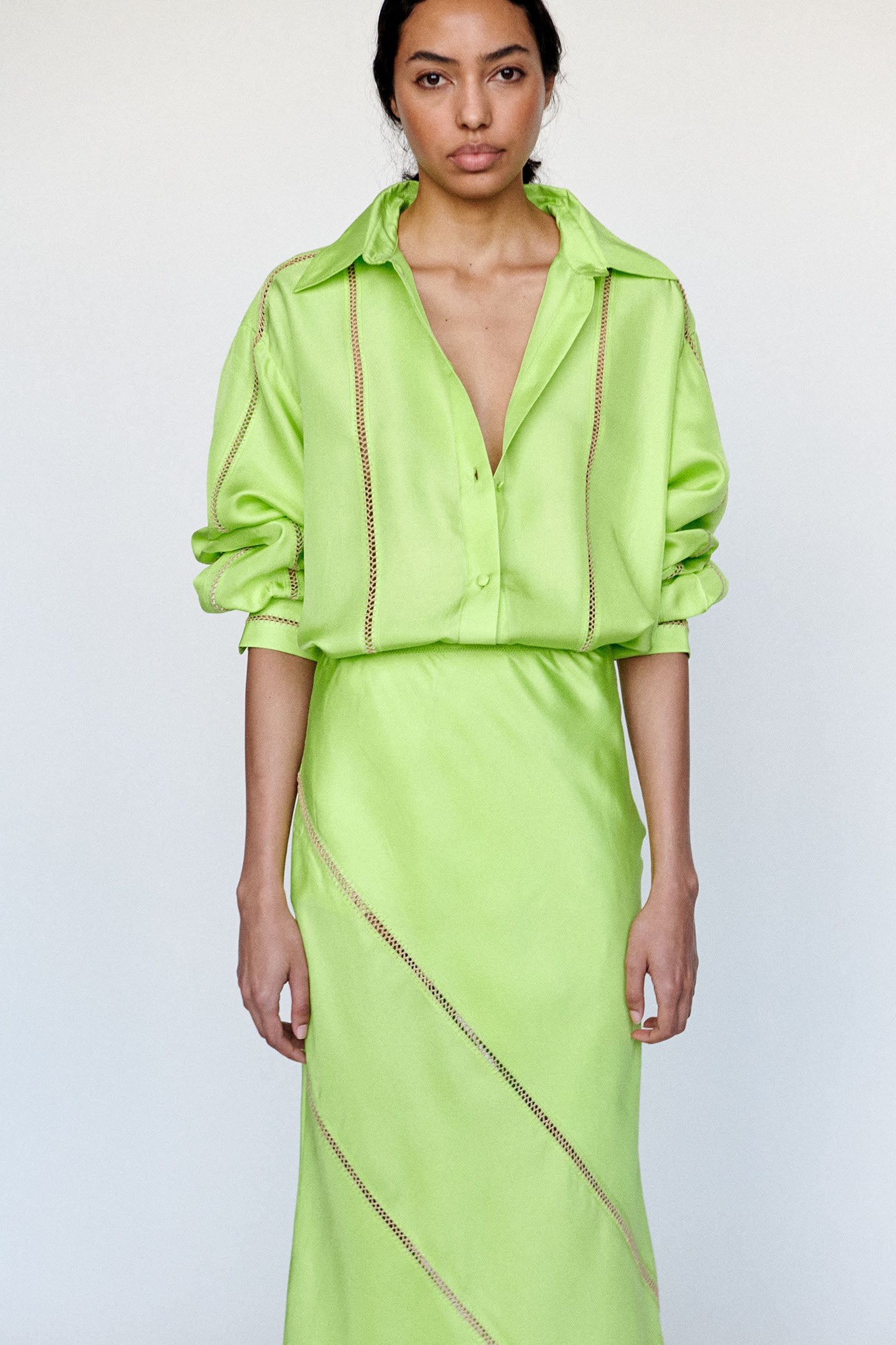 Model is wearing a bright green outfit with a matching button up and skirt with intricate detailing on both photographed on a white background