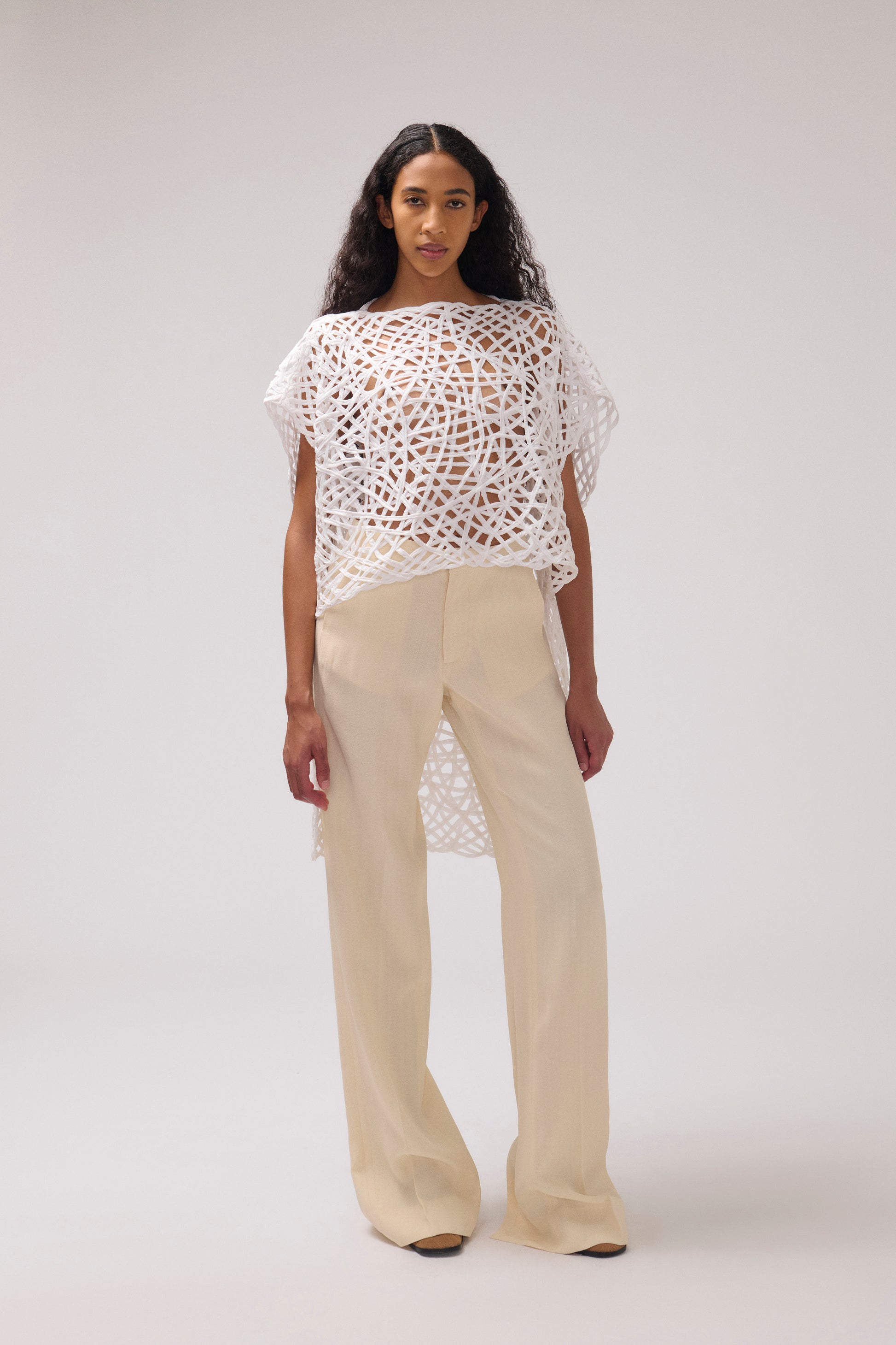 A model stands against a light gray studio background wearing a white open-knit cropped top with wide short sleeves and cream wide-leg trousers.