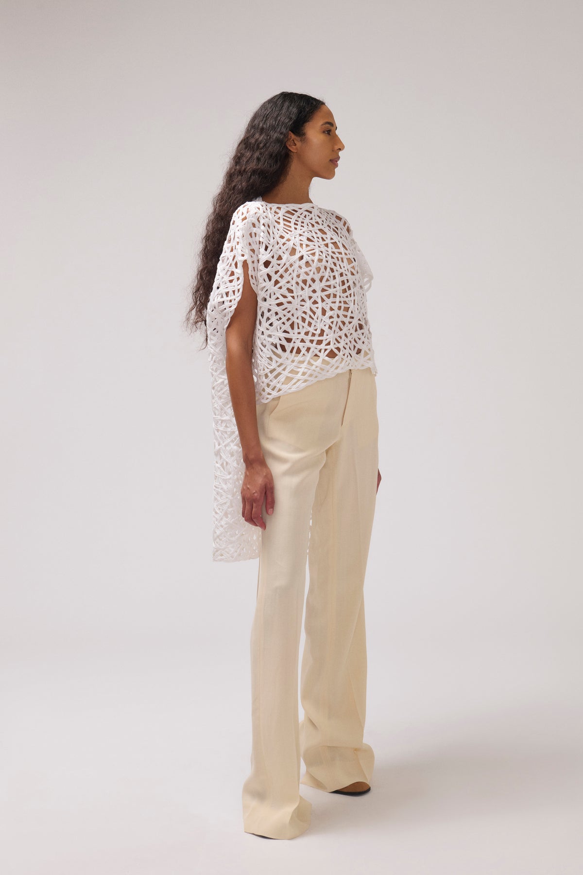A model is shown from the back wearing a white crochet top with draped sleeves and cream-colored wide-leg trousers, set against a plain light gray studio background.