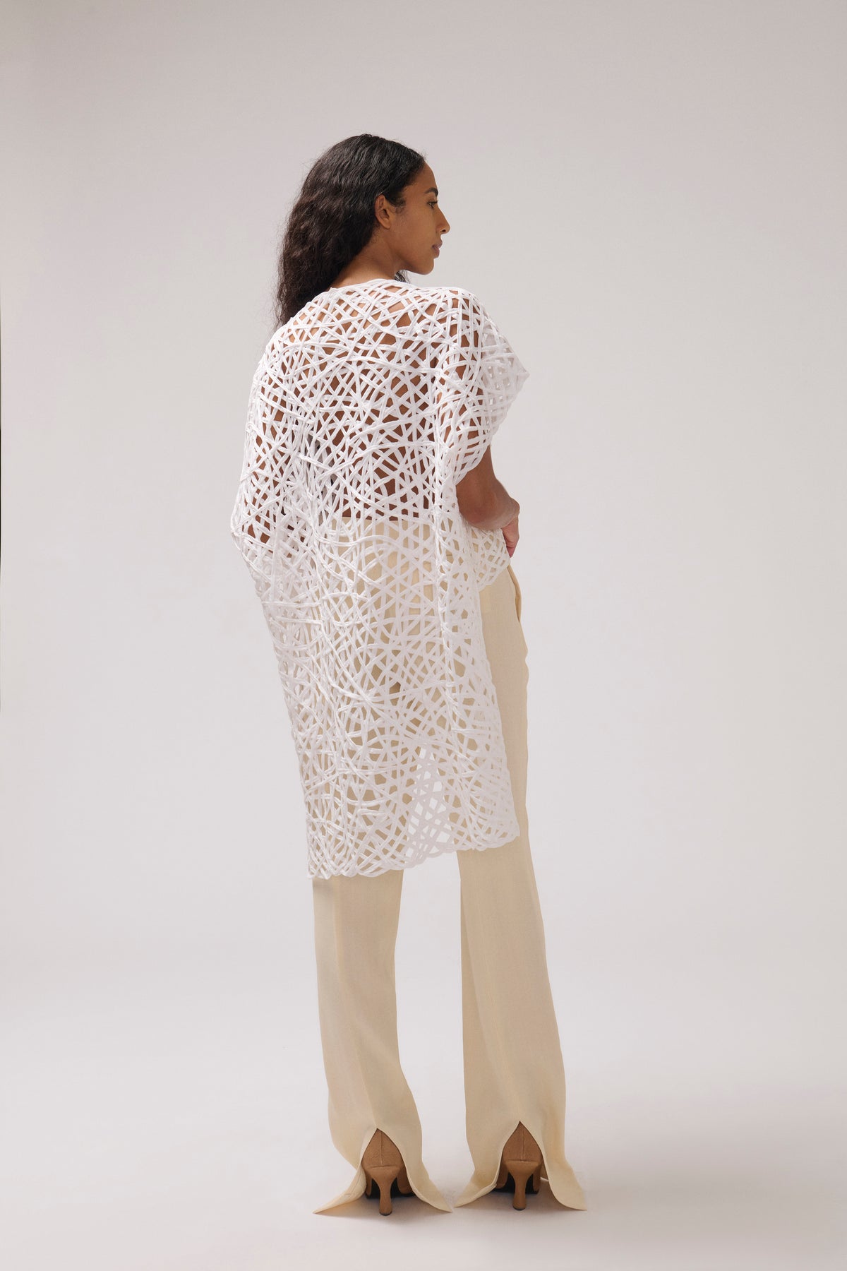 A woman shown from the back wearing a white open-knit crochet tunic top and cream wide-leg trousers against a light gray studio background.