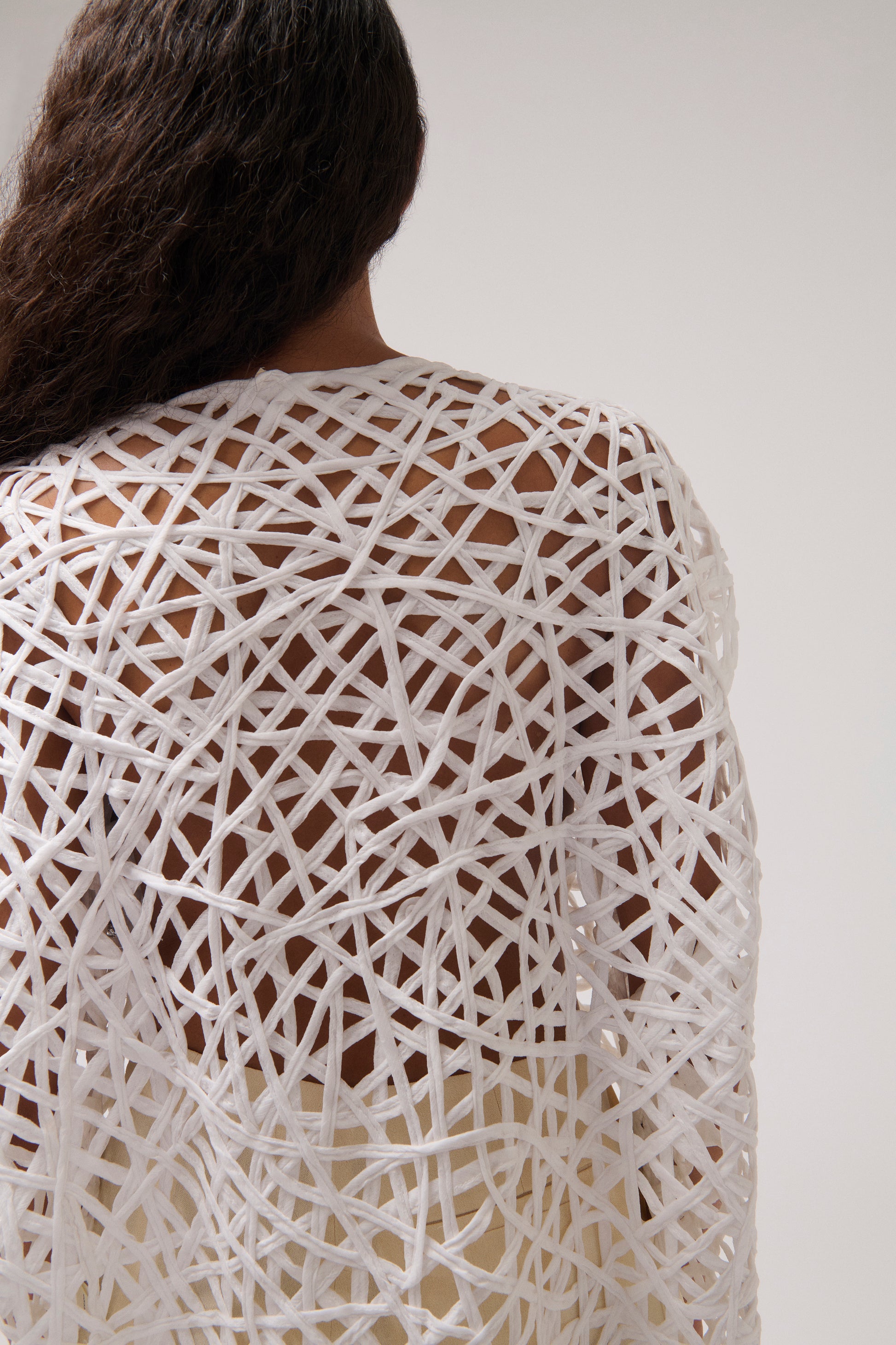 A close-up back view of a person wearing a white open-knit top with an intricate, woven geometric pattern over the skin.