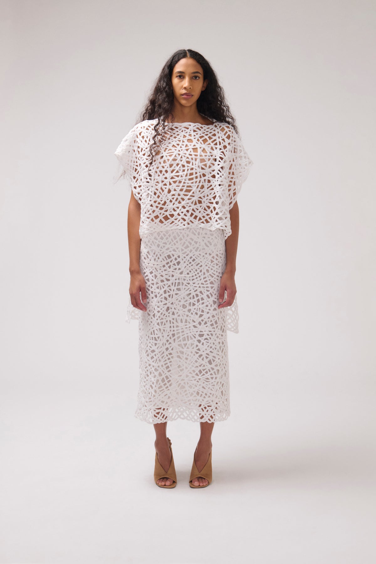 A model stands against a light gray studio background wearing a white crochet or lace two-piece outfit with a short flutter-sleeve top and a midi skirt, paired with tan heeled sandals.