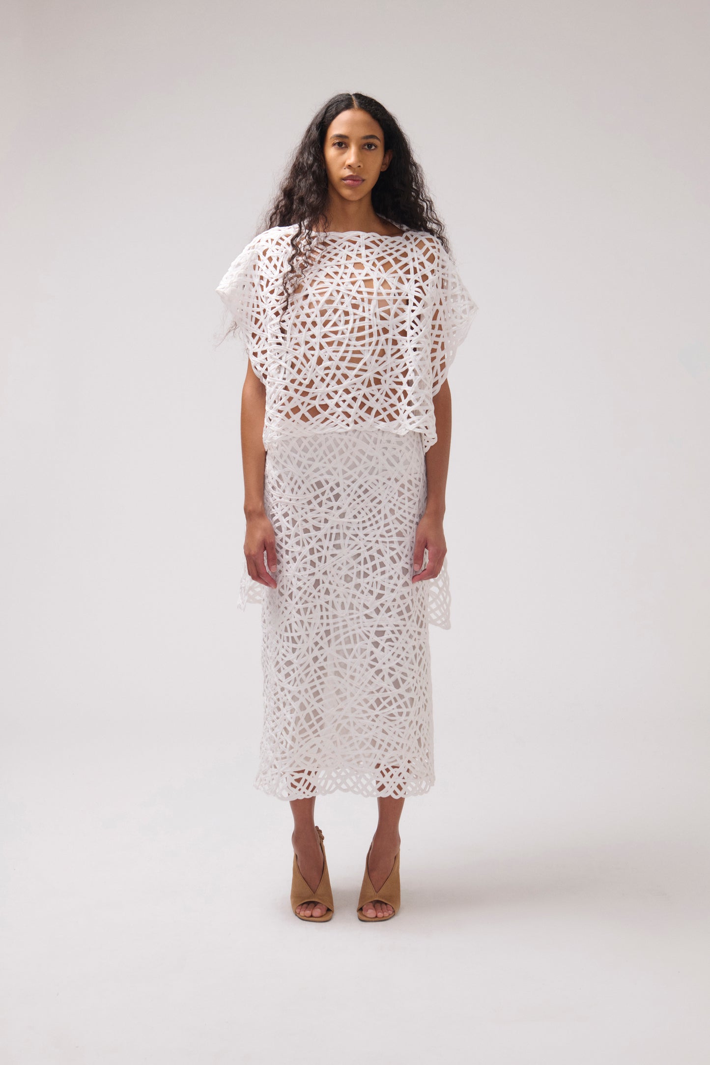 A model stands against a light gray studio background wearing a white crochet lace two-piece set consisting of a short-sleeve cropped top and a midi skirt, paired with tan high-heeled sandals.
