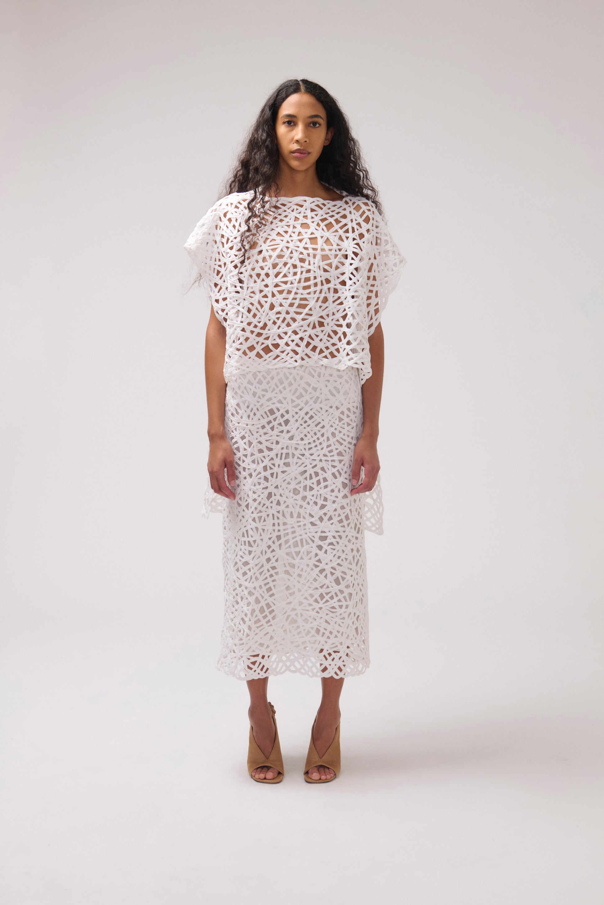 A model stands against a light gray studio background wearing a white crochet lace two-piece set consisting of a short-sleeve cropped top and a midi skirt, paired with tan high-heeled sandals.