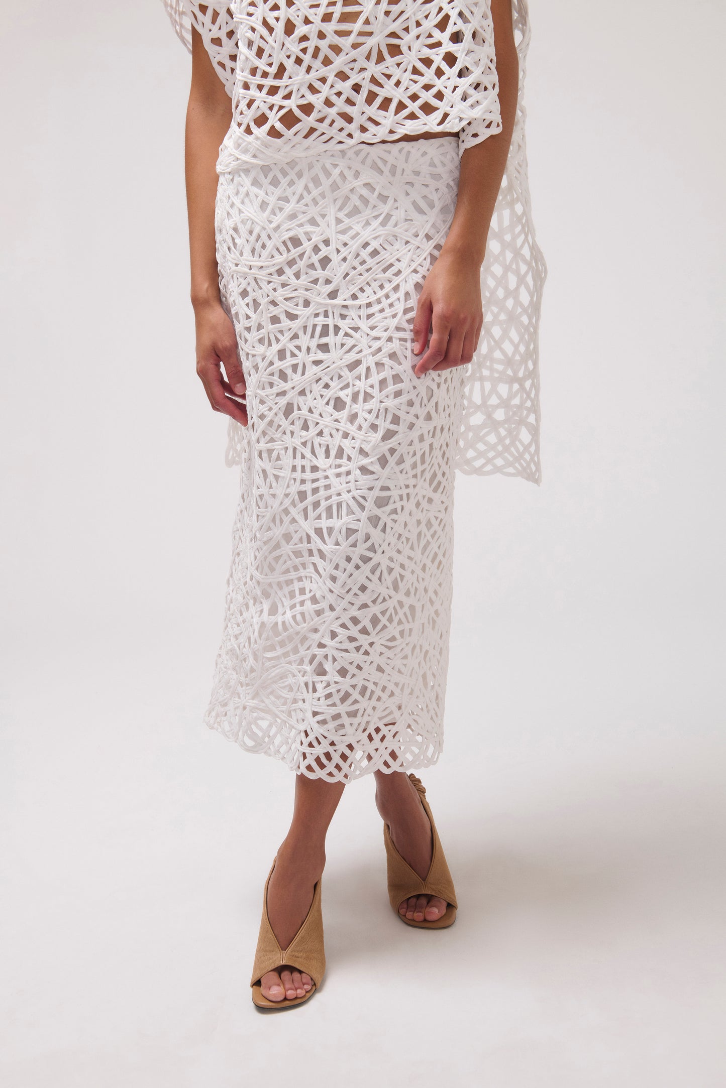 A model wearing a white crochet midi skirt with a matching crochet top and a sheer white wrap, paired with tan heeled sandals, against a plain white studio background.