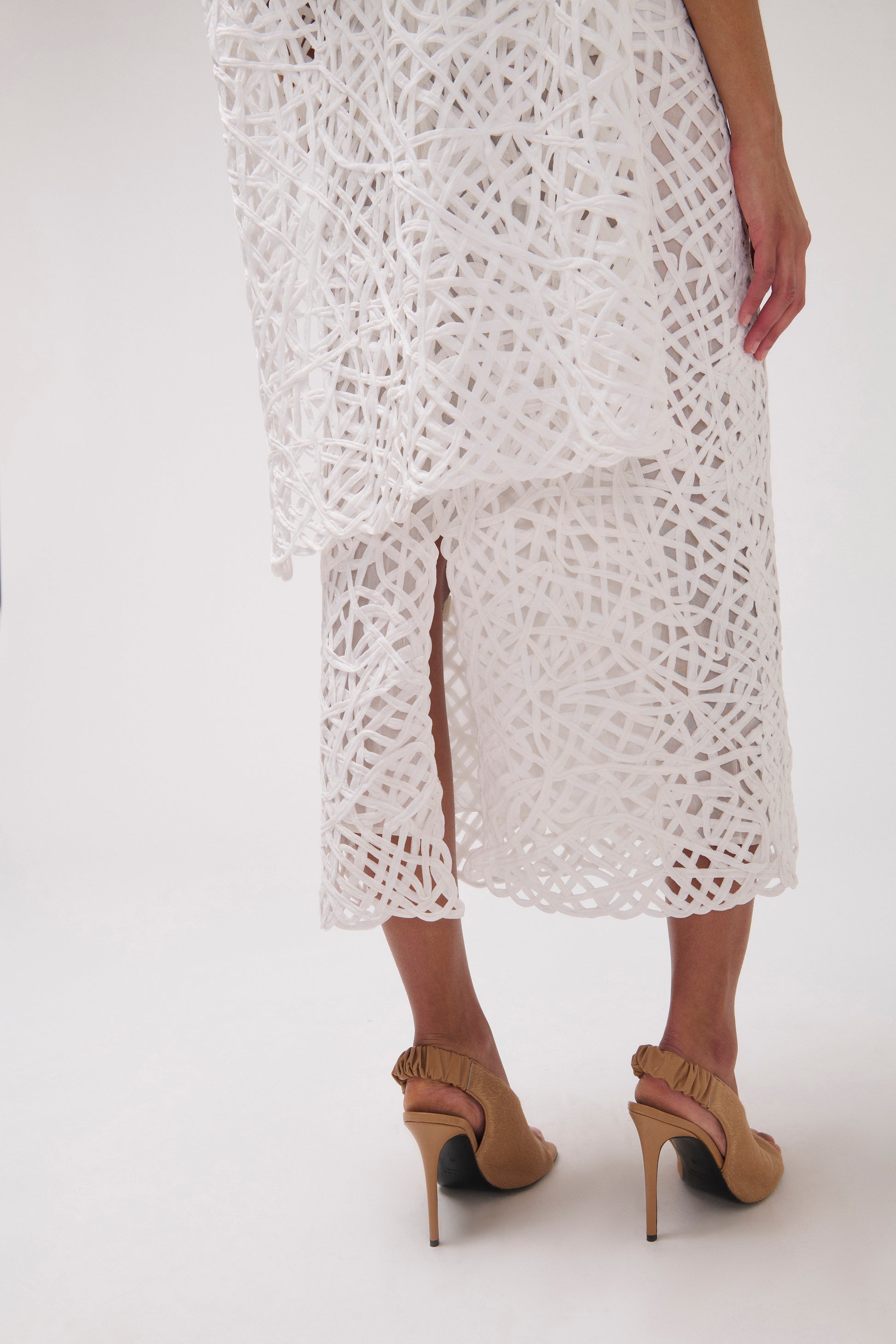 White crochet midi skirt with a side slit, shown on a model from the back wearing beige slingback high heels against a white studio background.