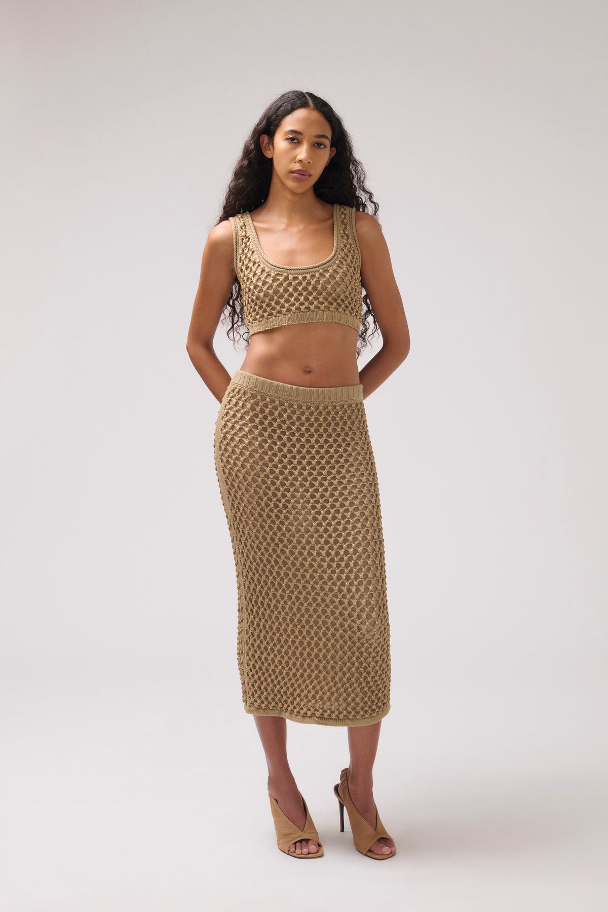 A model stands against a light gray studio background wearing a matching beige open-knit two-piece set consisting of a cropped tank top and a midi skirt, paired with beige heeled sandals.