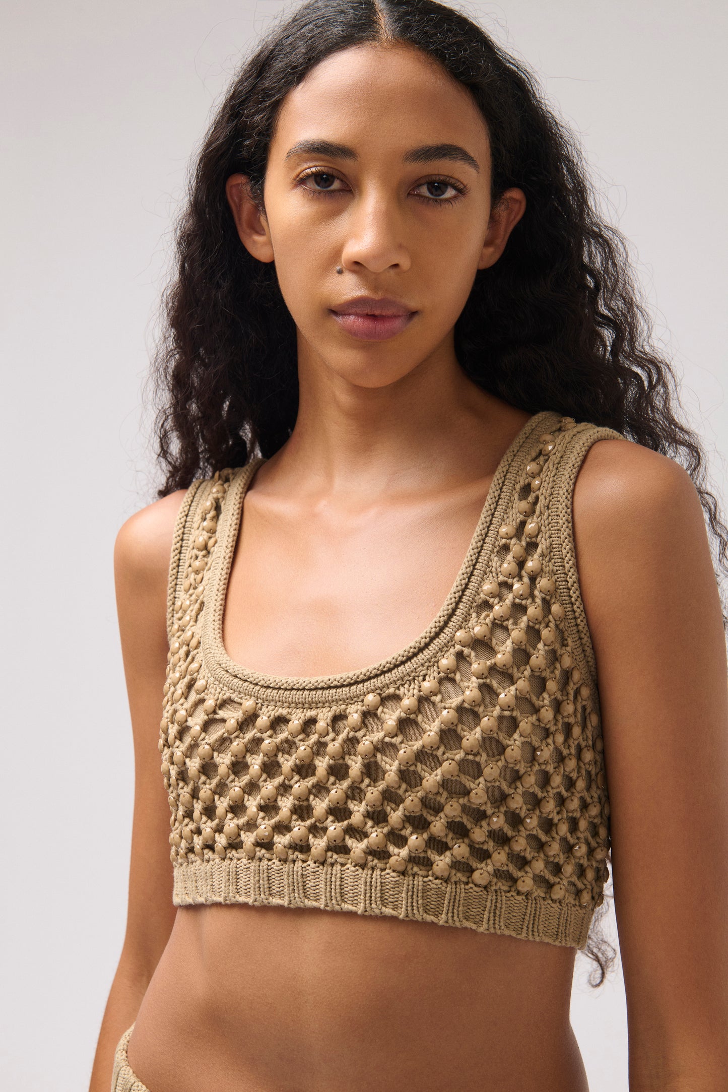 A woman wearing a beige crochet knit crop top with a scoop neckline, photographed against a neutral light gray studio background.