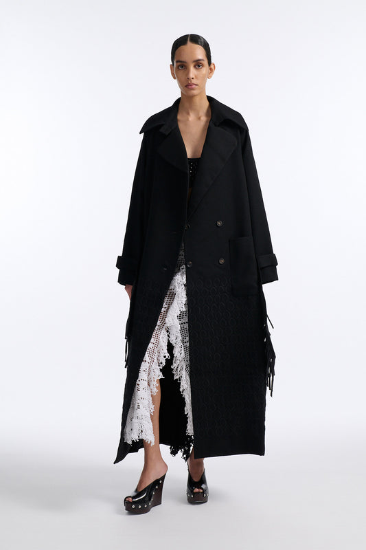 Female model with slicked back black hair in a bun wears long black wool coat with oversized collar and fringe belt.