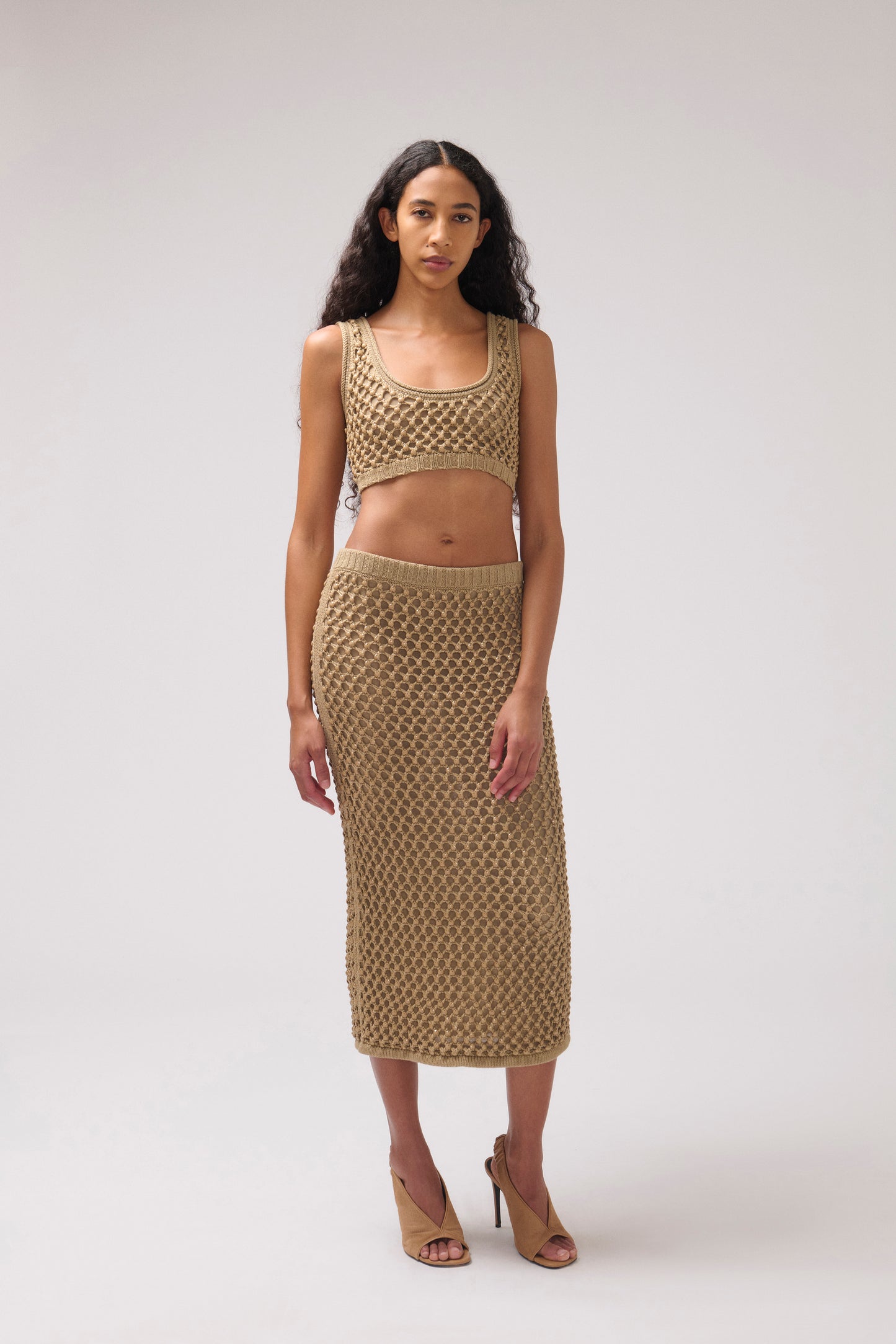A model stands against a light gray studio background wearing a matching beige knit two-piece set consisting of a cropped tank top and a midi skirt, paired with tan heeled sandals.