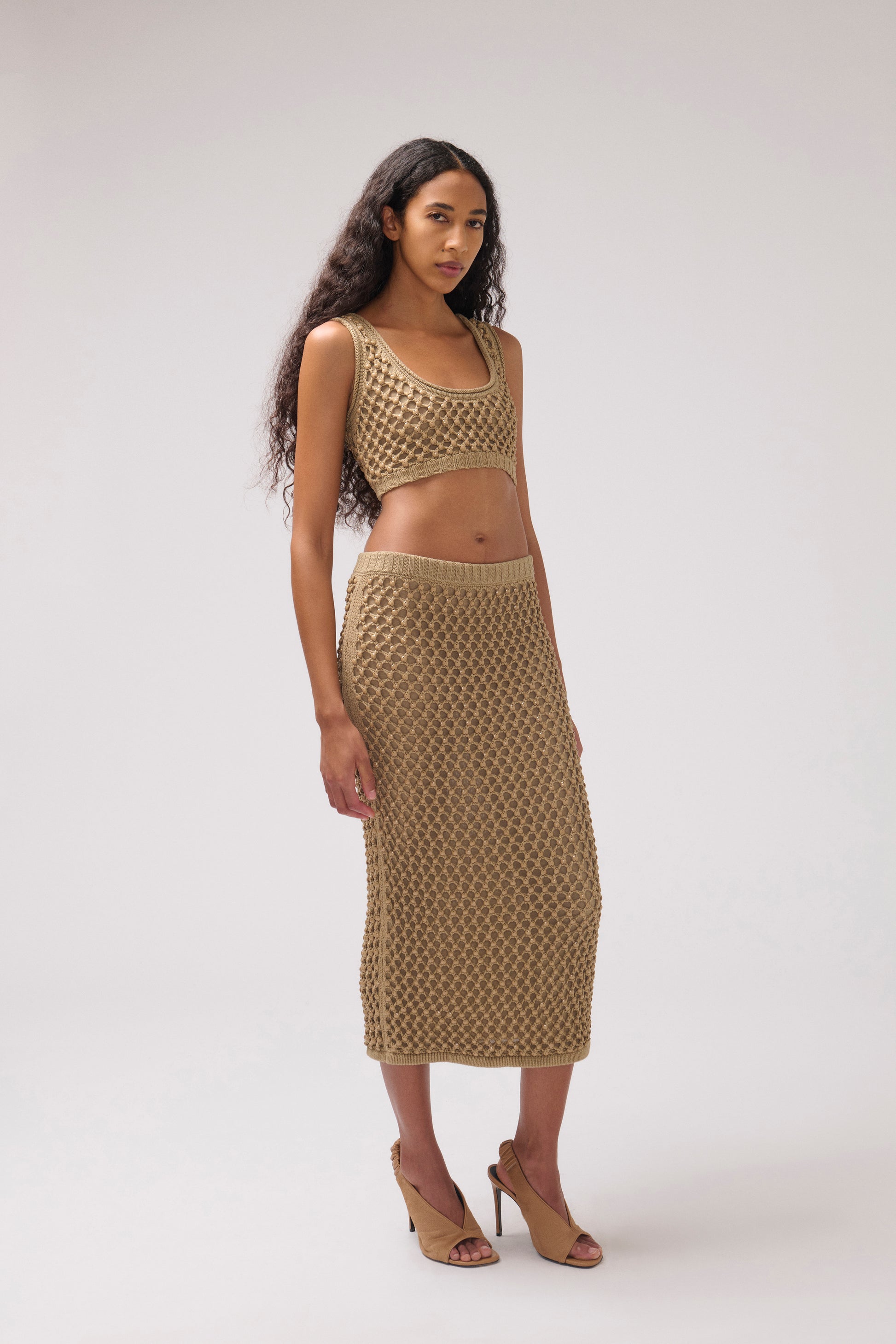 A model wearing a matching beige crochet knit two-piece set consisting of a cropped tank top and a midi skirt, paired with tan high-heeled sandals, against a light gray studio background.