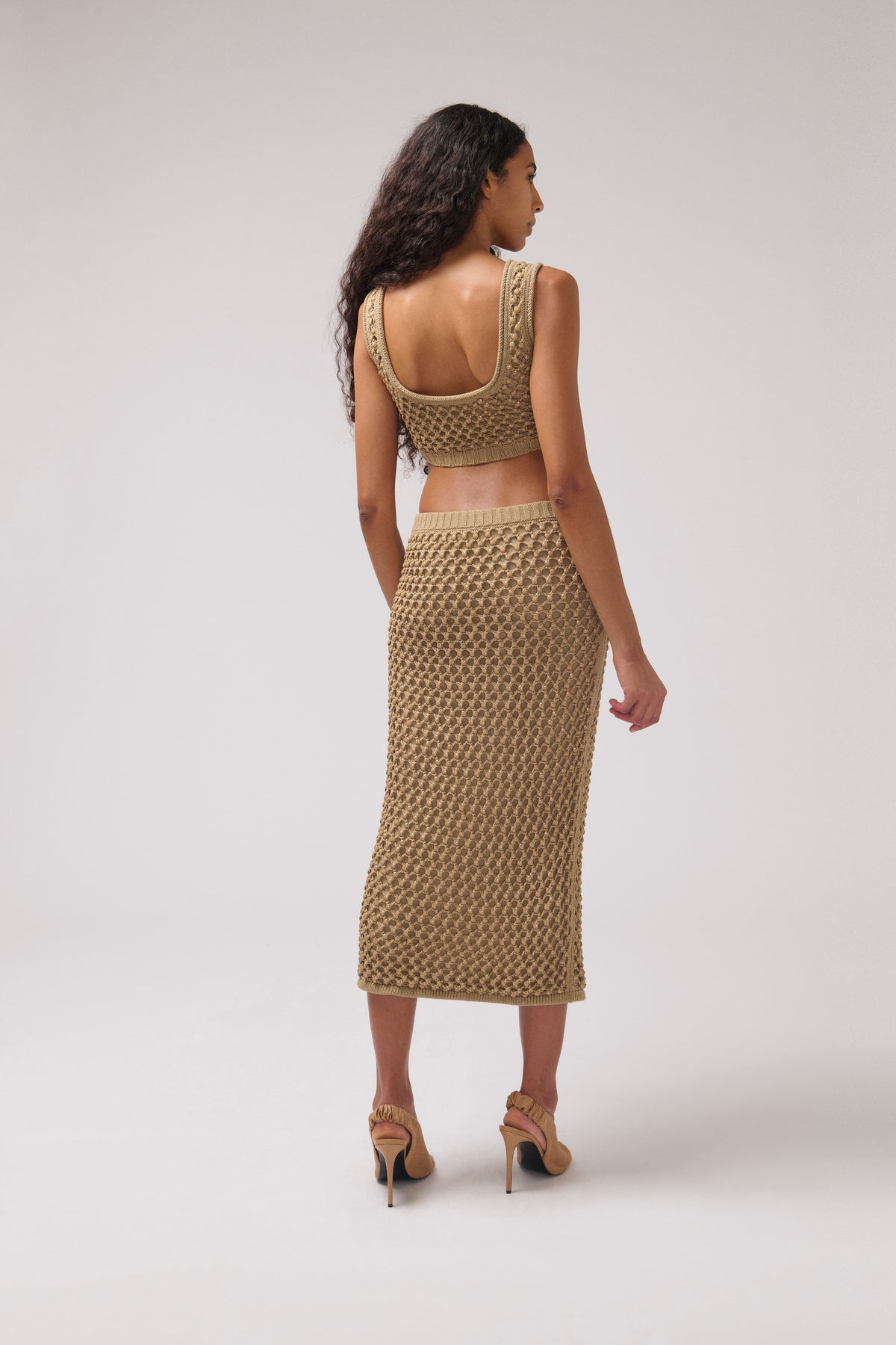A woman shown from the back wearing a matching textured knit two-piece set consisting of a sleeveless crop top and a midi skirt in a warm beige color, paired with tan high heels, against a light gray studio background.