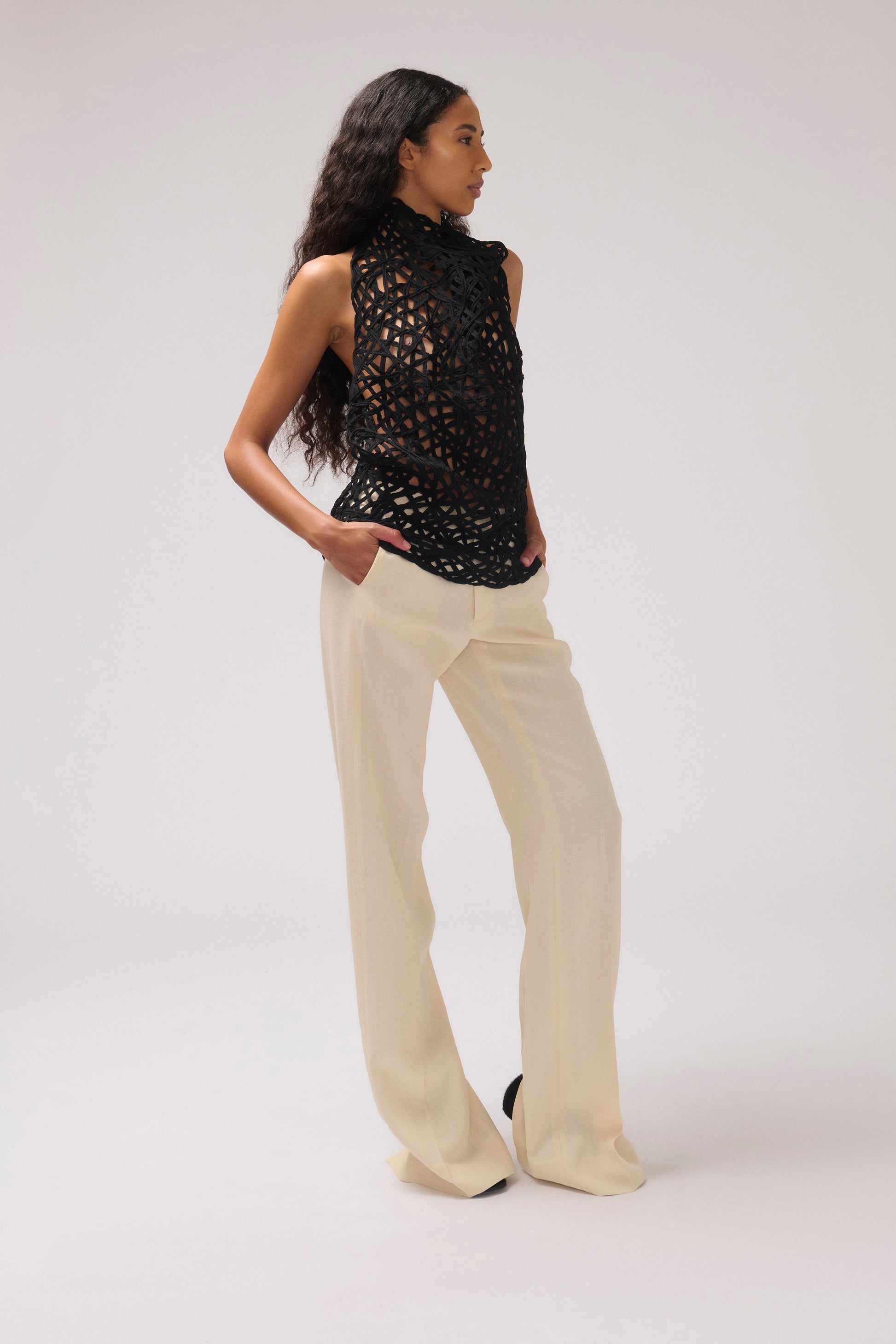A model is shown from the side wearing a sleeveless black crochet knit mock-neck top and cream wide-leg trousers, standing against a plain light gray studio background.