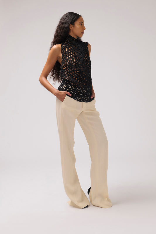 A model is shown from the side wearing a sleeveless black crochet knit mock-neck top and cream wide-leg trousers, standing against a plain light gray studio background.