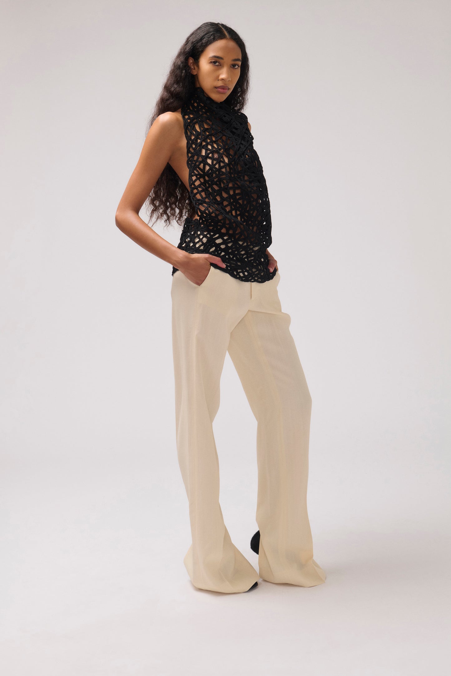 A model stands in a studio wearing a sleeveless black crochet top and cream wide-leg trousers against a light gray background.