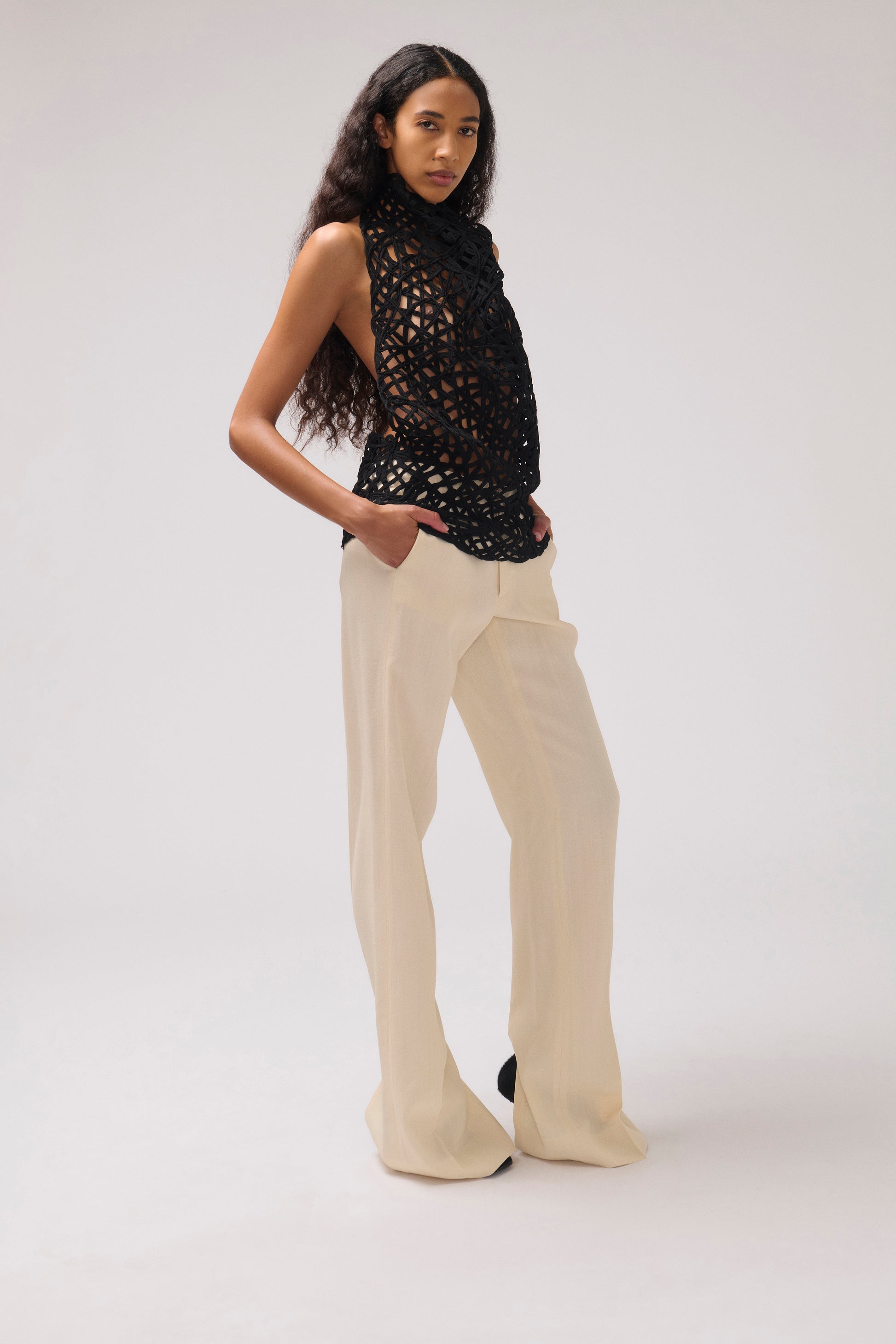 A model stands in a studio wearing a sleeveless black crochet top and cream wide-leg trousers against a light gray background.