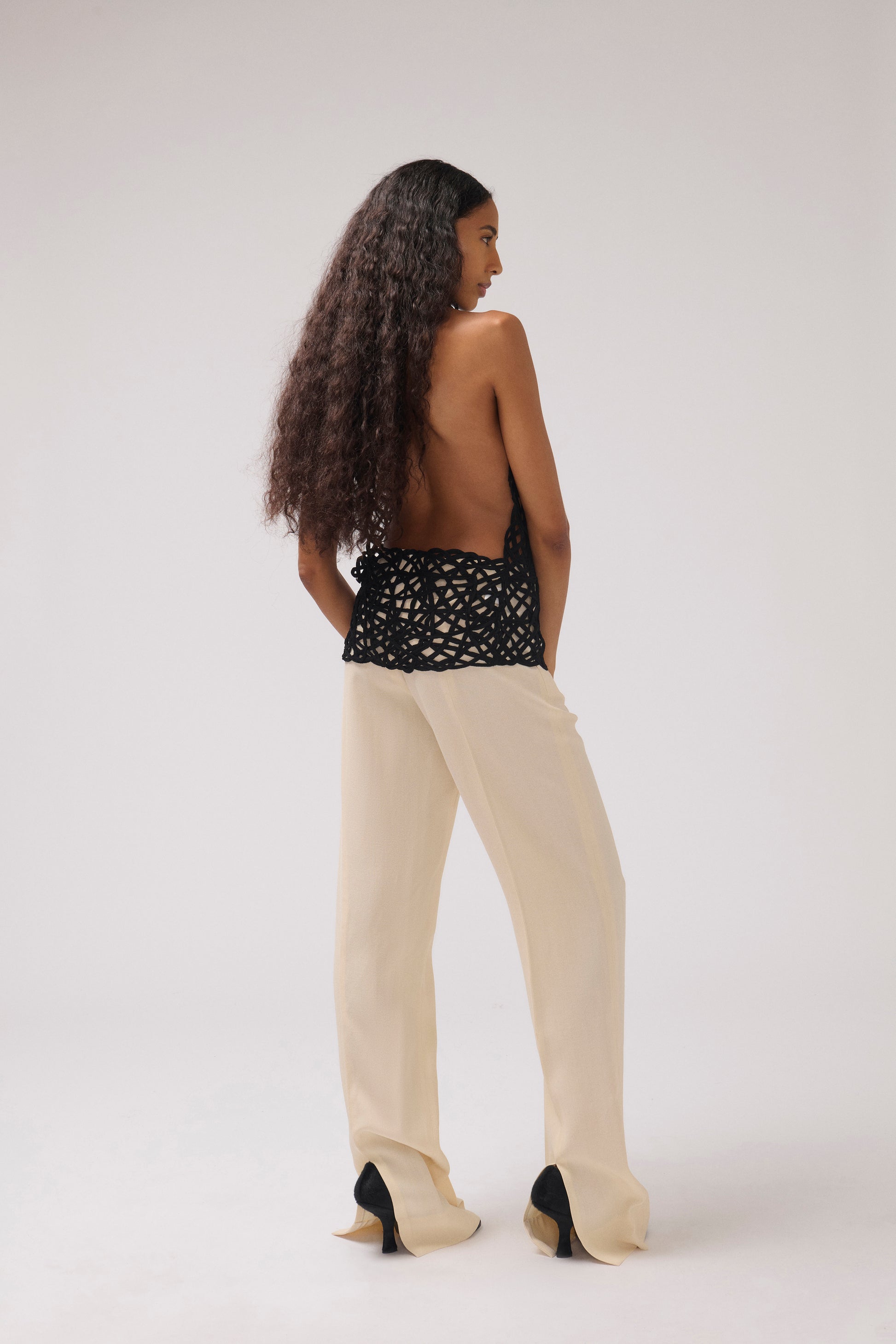 A model is shown from behind wearing a black crochet halter top with a wide back, cream wide-leg trousers, and black high-heeled shoes against a plain light gray studio background.