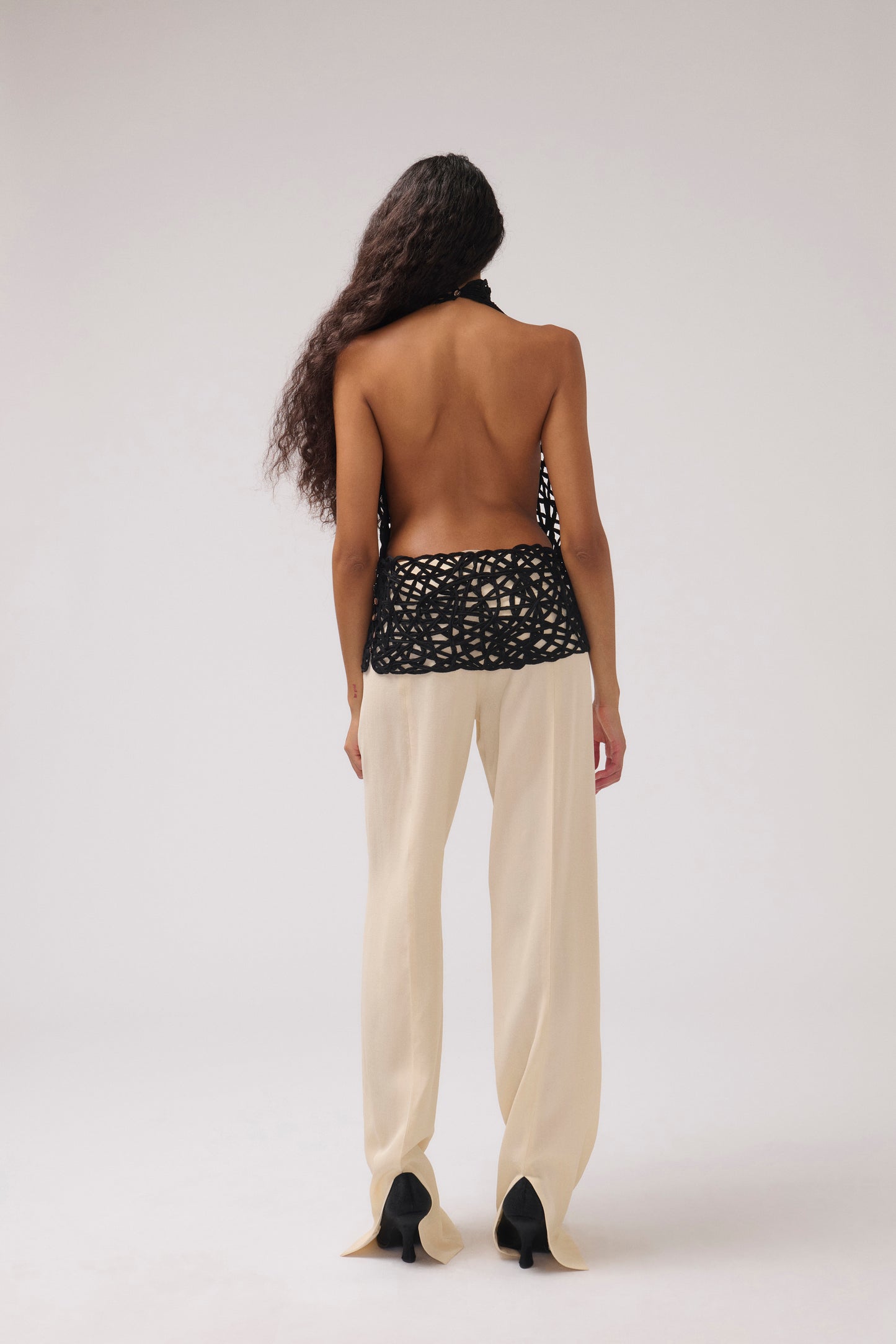 A model is shown from behind wearing a black crochet halter top with a wide waistband and cream wide-leg trousers, set against a plain light gray studio background.