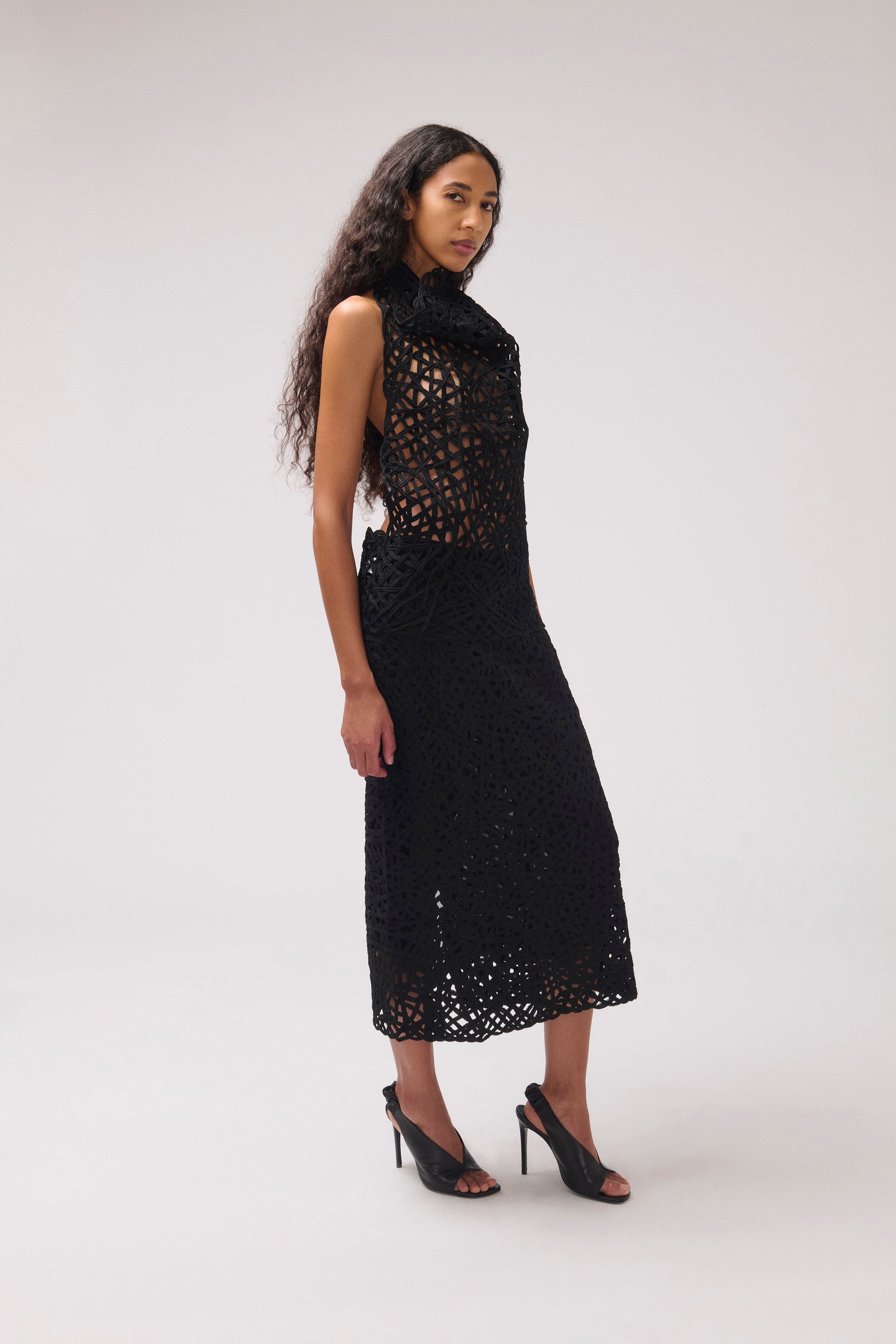 A model wearing a sleeveless black crochet midi dress with a high mock neck and black high-heeled sandals, standing against a light gray studio background.