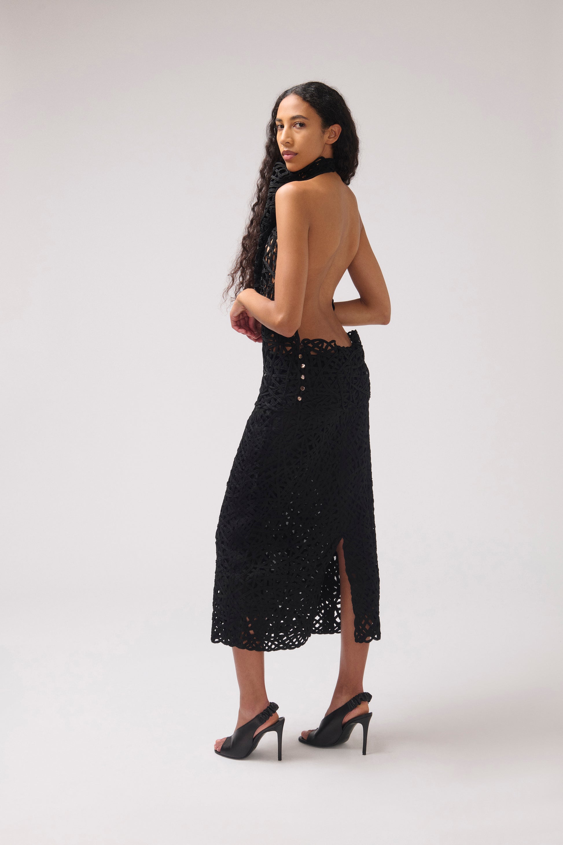 A woman modeling a black open-back midi dress with a high slit and button details, paired with black high-heeled sandals, photographed against a light gray studio background.
