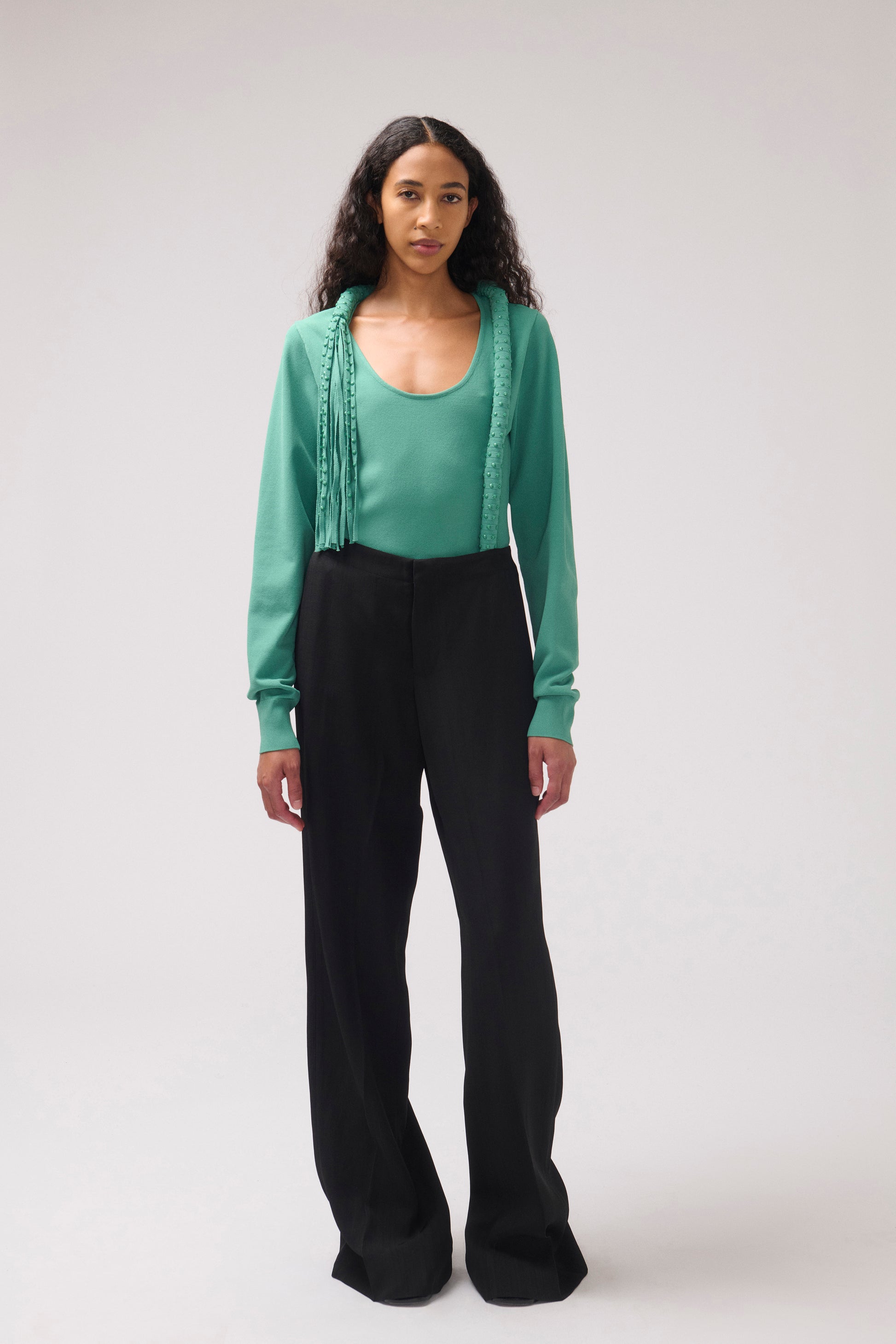 A model stands against a light gray studio background wearing a mint green long-sleeve top with fringe detailing, a matching scoop-neck bodysuit, and black wide-leg trousers.