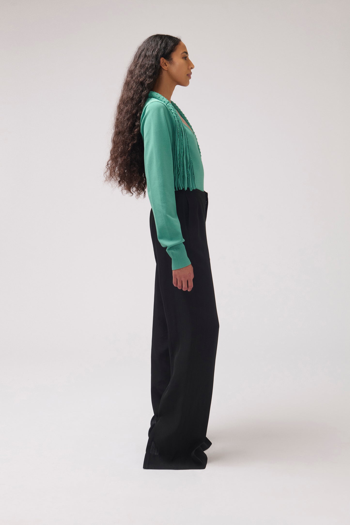 A woman stands in profile wearing a long-sleeve green top with a fringed neckline and black wide-leg pants against a plain light gray studio background.