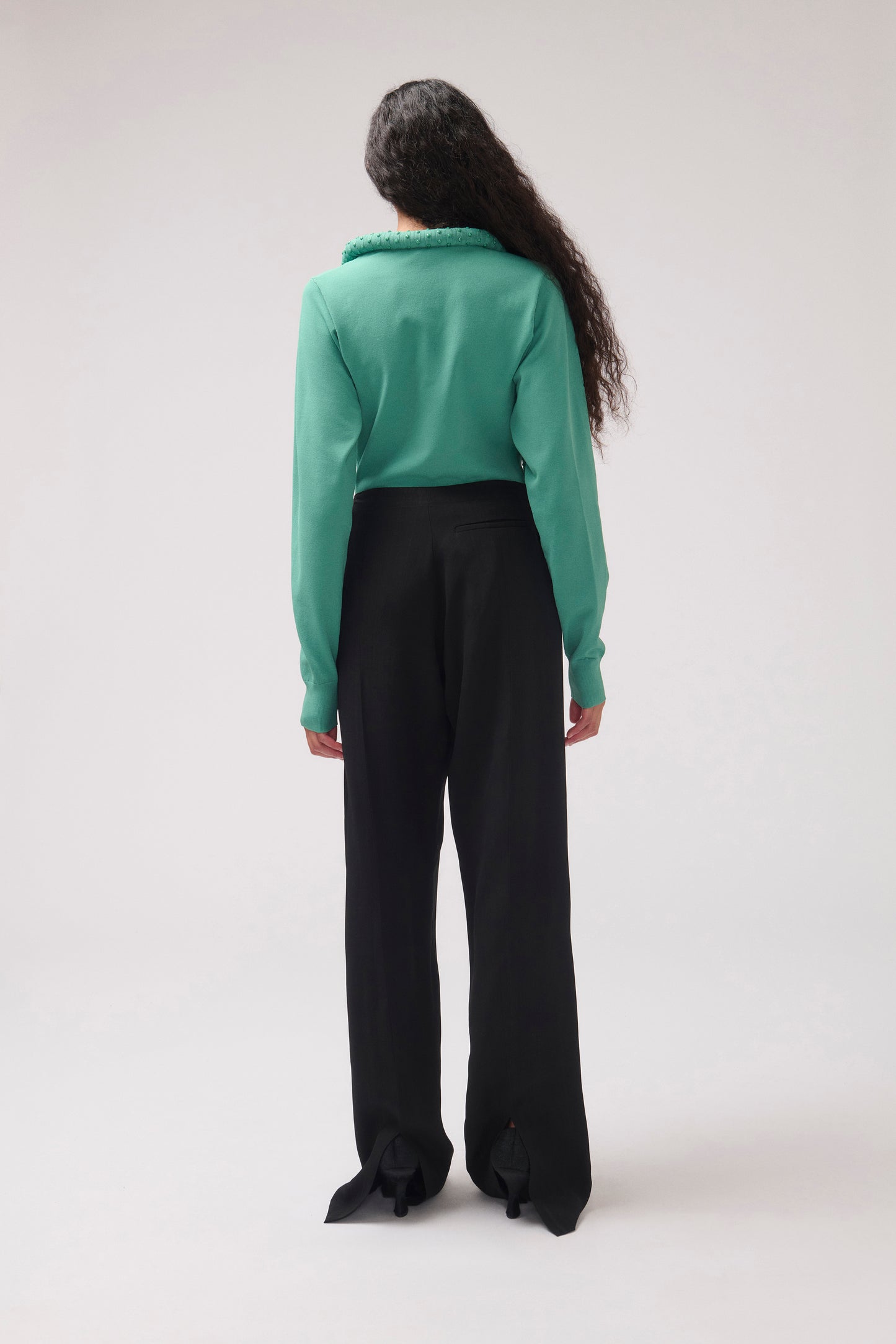 A model viewed from the back wearing a mint green long-sleeve top with a fuzzy collar and black wide-leg trousers, set against a plain light gray studio background.