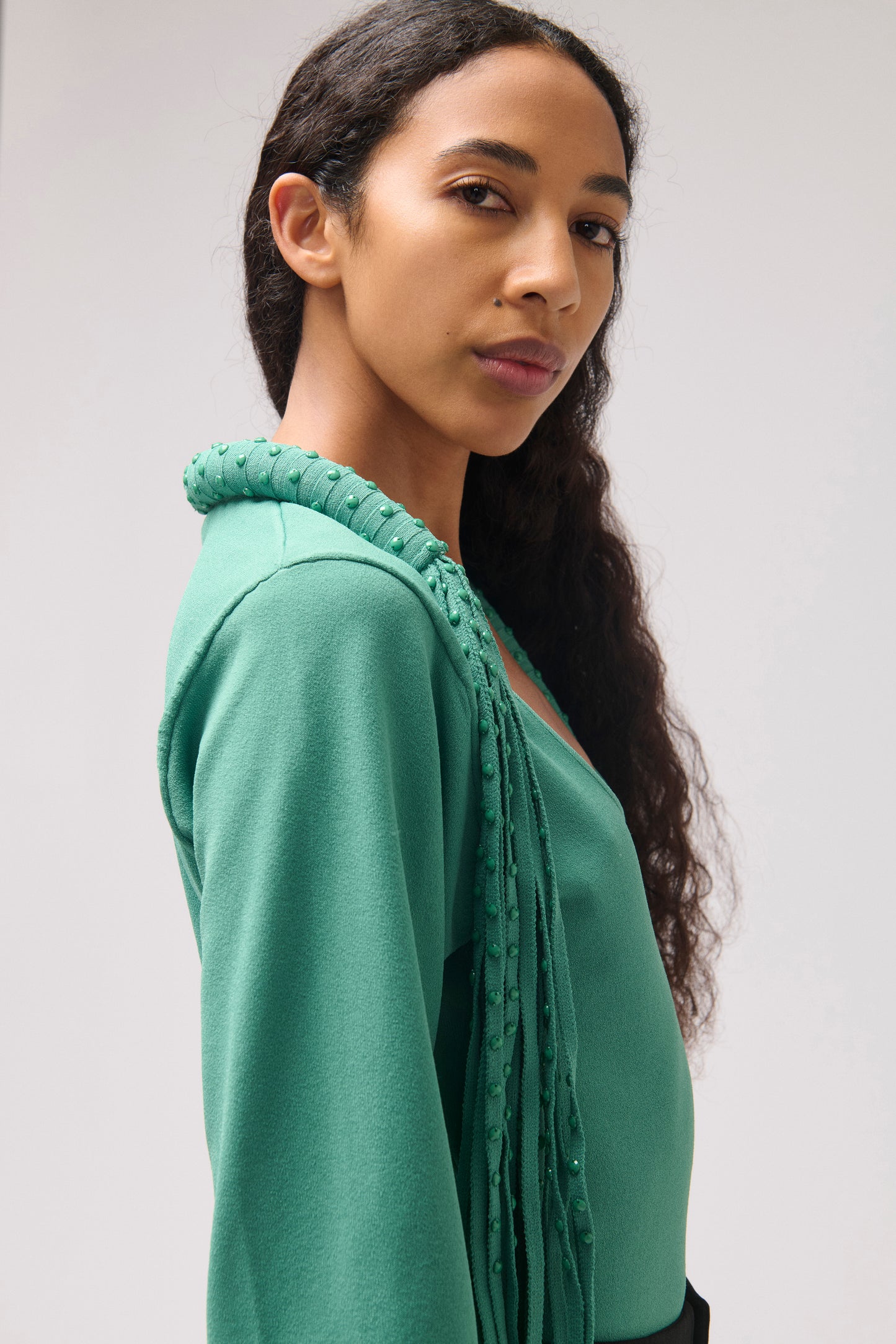 A woman wearing a teal green knit top with a textured, raised collar and draped fringe details, shown in a close-up profile shot against a light gray background.