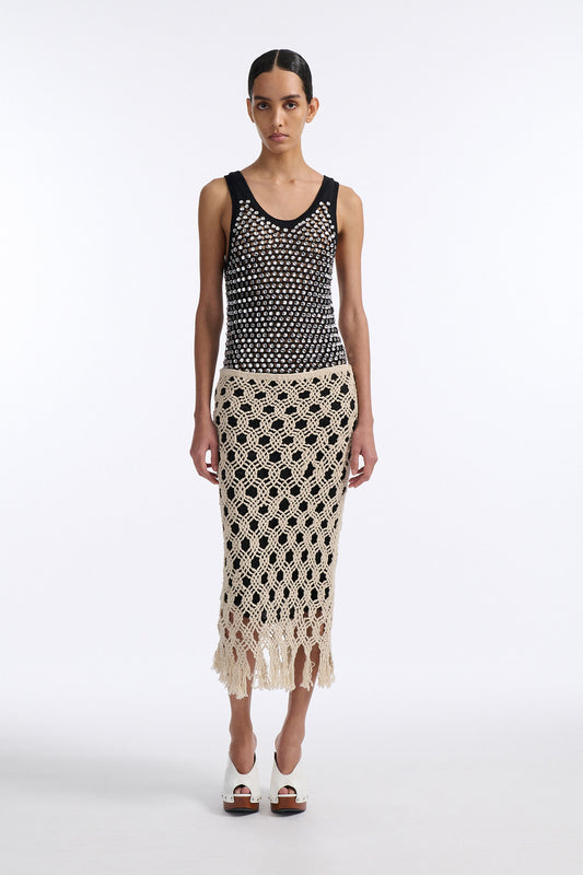 Female model with slicked back black hair in a bun wears black chunky mesh tank top with clear hand-sewn crystals and a cream colored macrame skirt with a black lining.