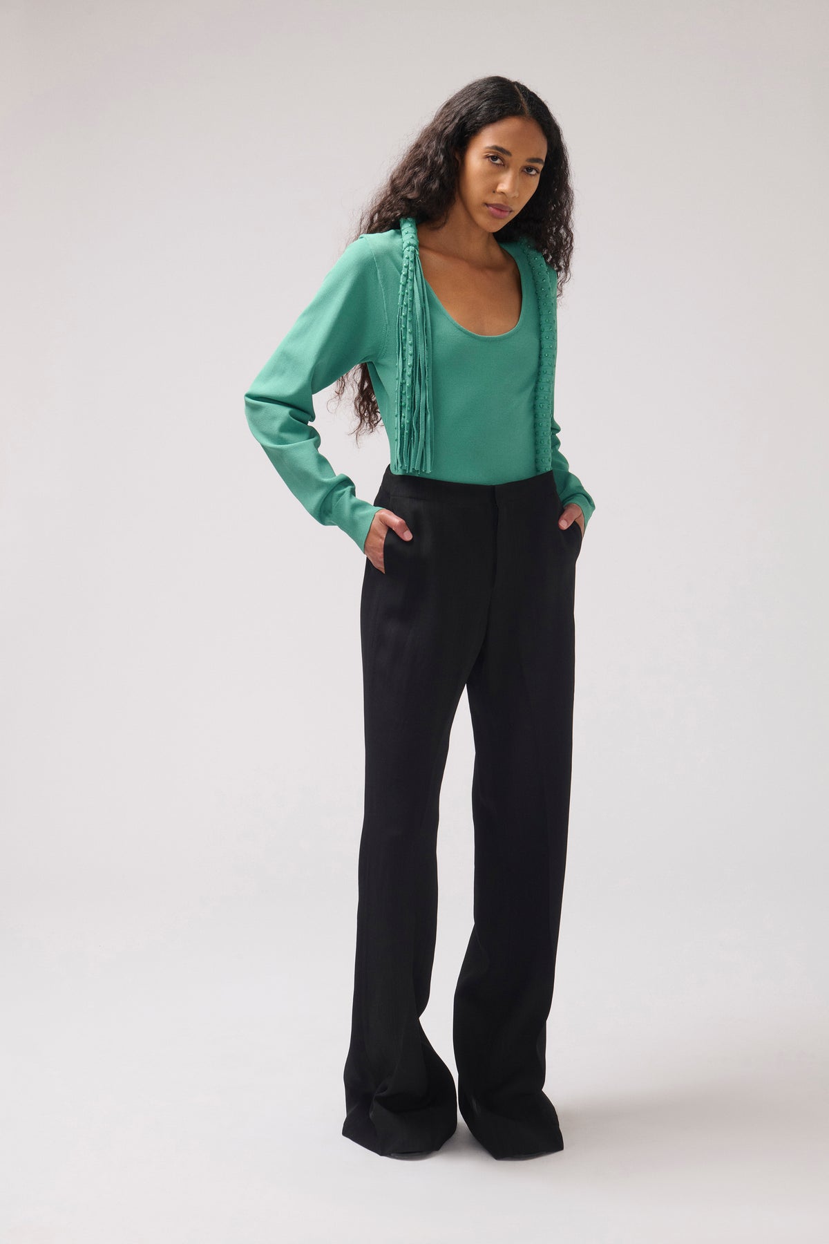 A model stands against a light gray studio background wearing a green long-sleeve top with decorative trim and black wide-leg trousers.
