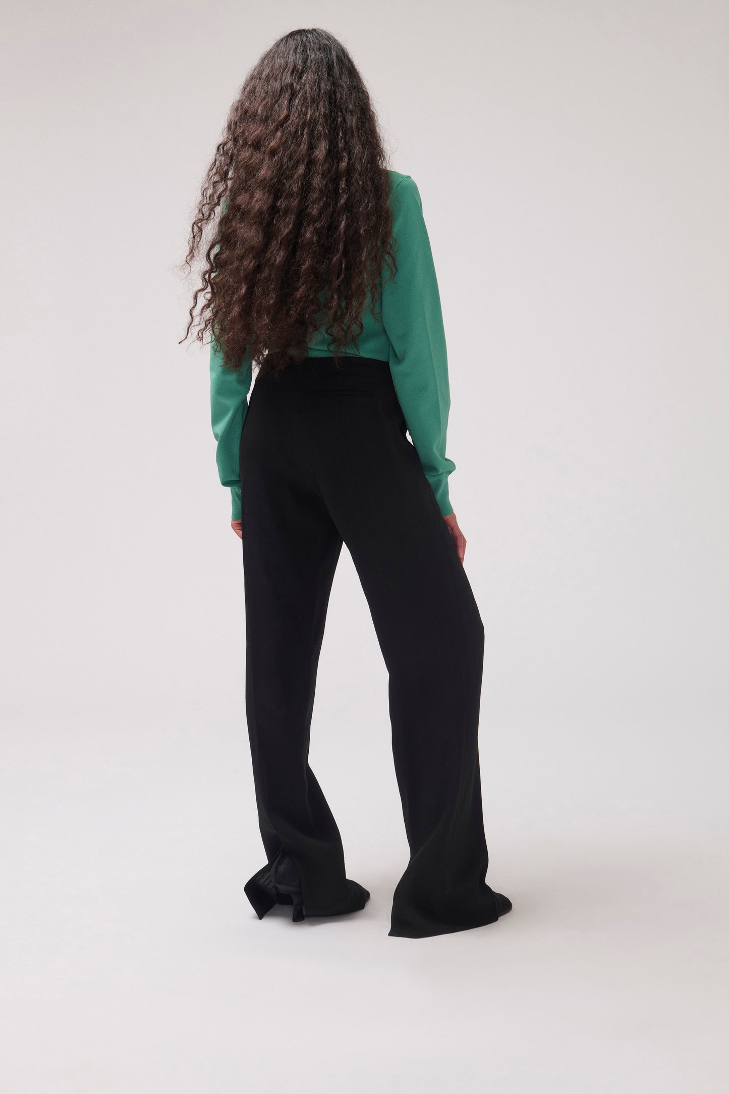 A model viewed from behind wearing black wide-leg trousers and a green long-sleeve top against a plain light gray studio background.