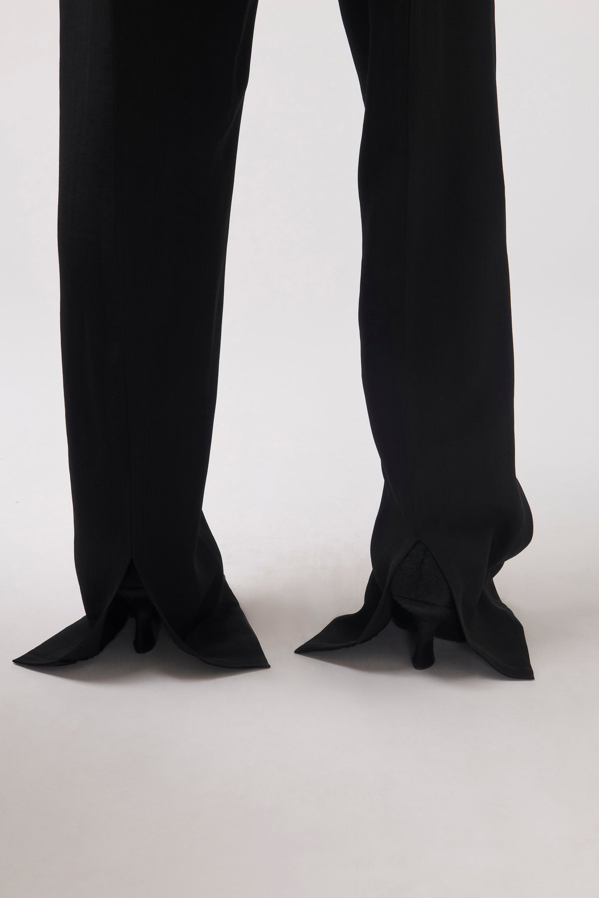 Black wide-leg trousers shown from the back, featuring dramatic pointed flared hems and kitten heels, photographed against a clean light gray studio background.