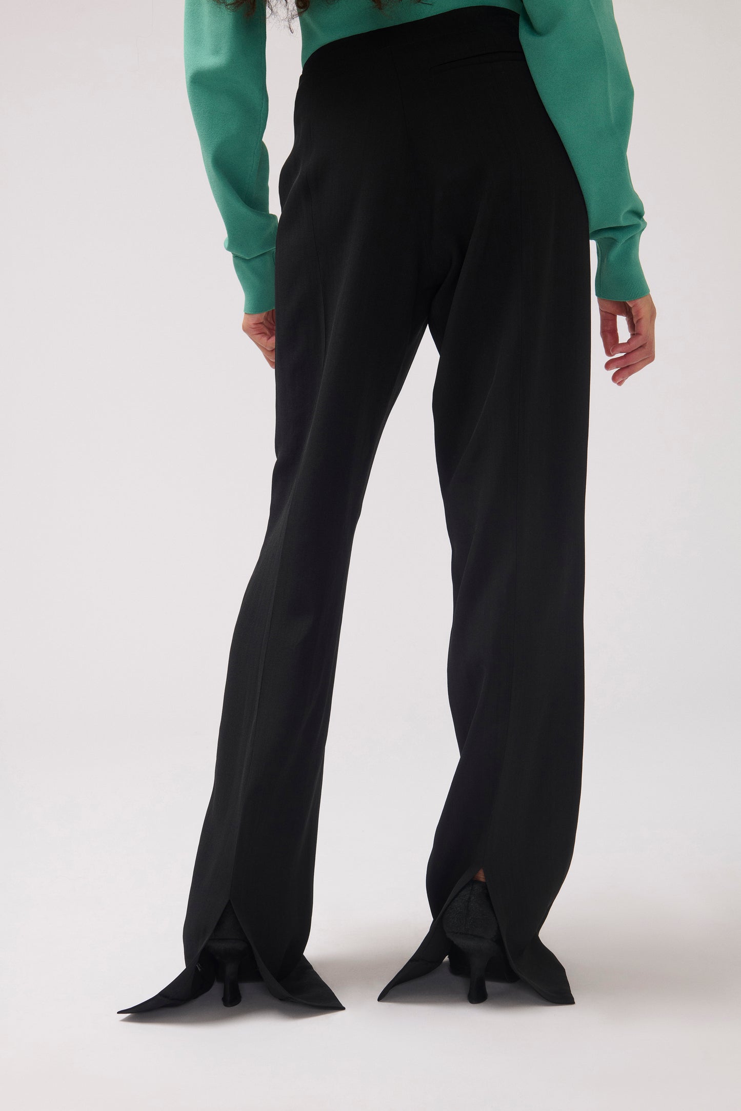 A model is shown from the back wearing black wide-leg trousers with unique pointed flared hems and a green long-sleeve top, photographed against a light gray studio background.