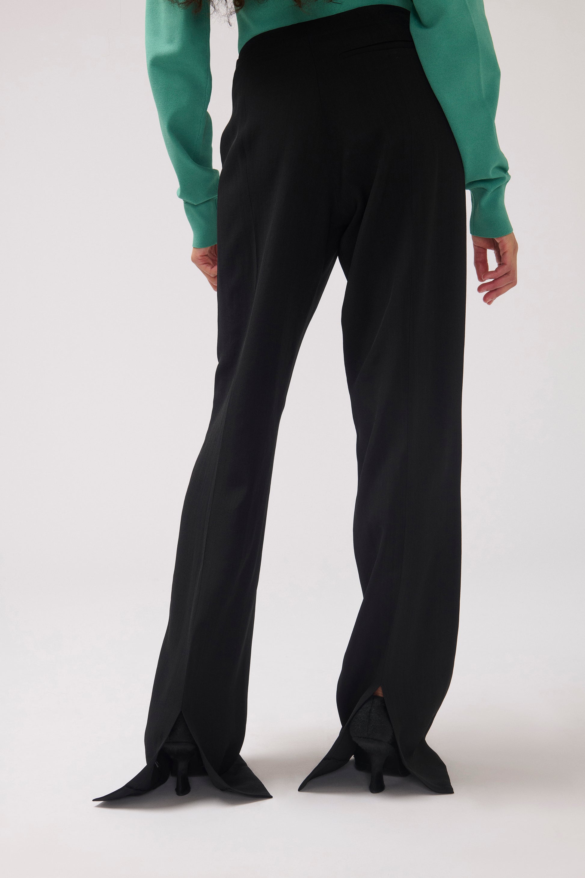A model is shown from the back wearing black wide-leg trousers with unique pointed flared hems and a green long-sleeve top, photographed against a light gray studio background.