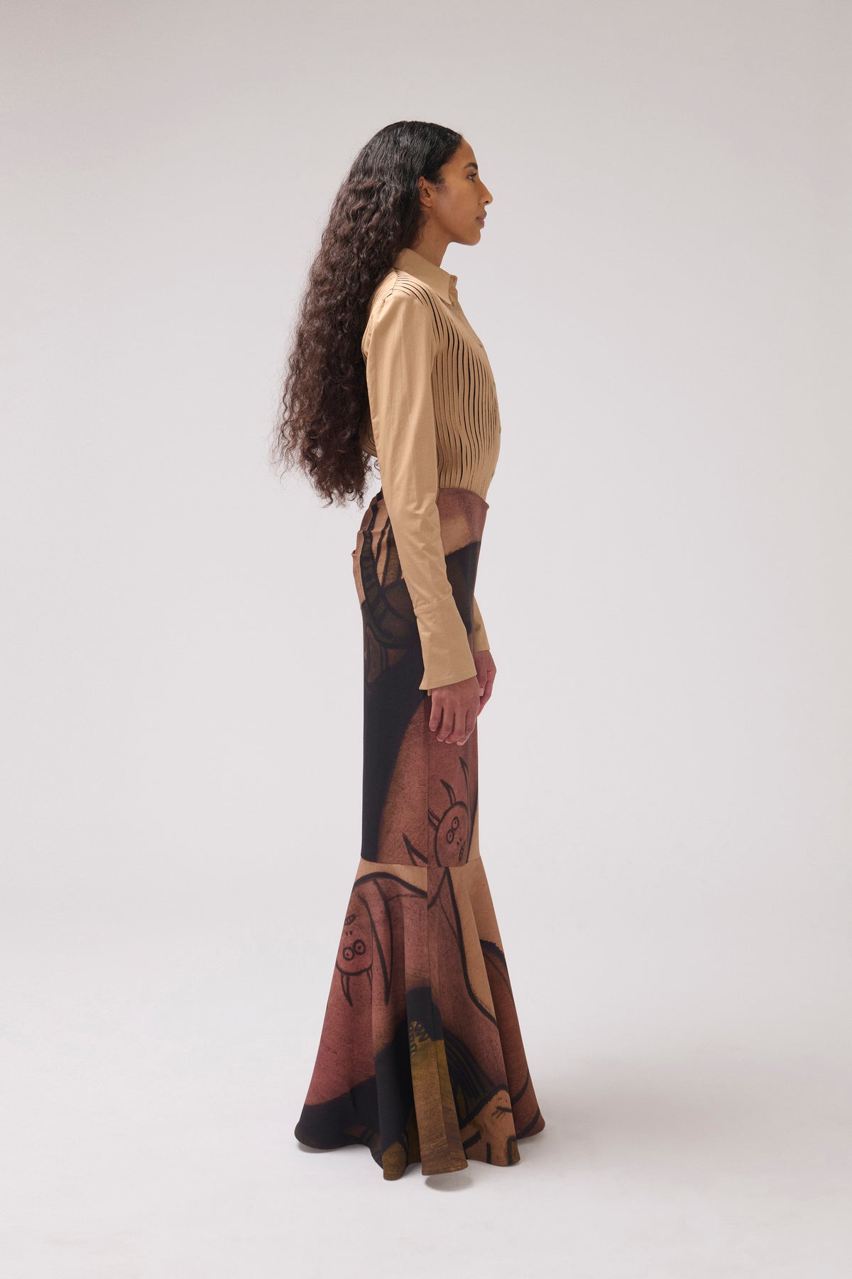 A model is shown in profile wearing a beige long-sleeve crop top and a high-waisted, floor-length skirt with a brown and black abstract floral print, set against a plain light gray studio background.