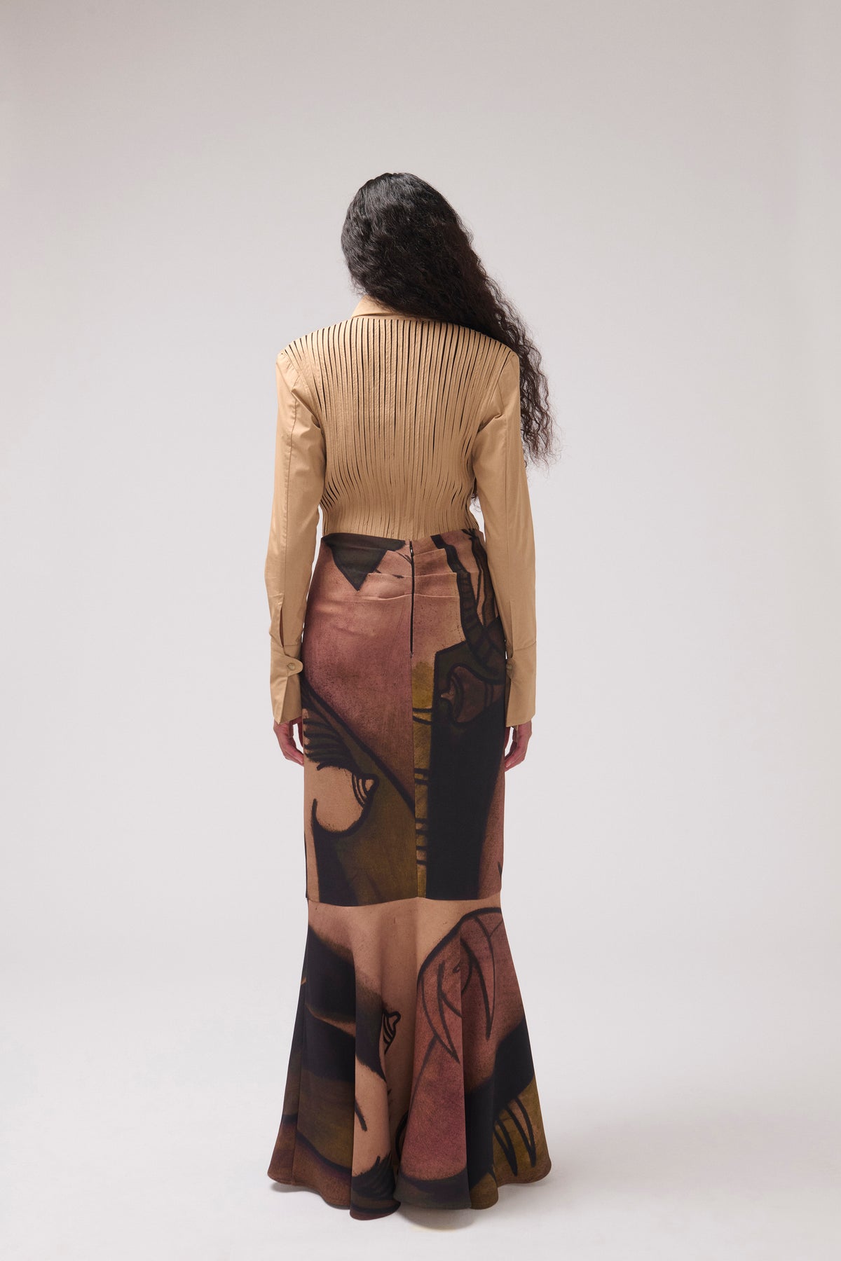 A model viewed from behind wearing a beige long-sleeve pleated top and a matching long skirt with a large abstract portrait print in beige, brown, and black, against a light gray studio background.