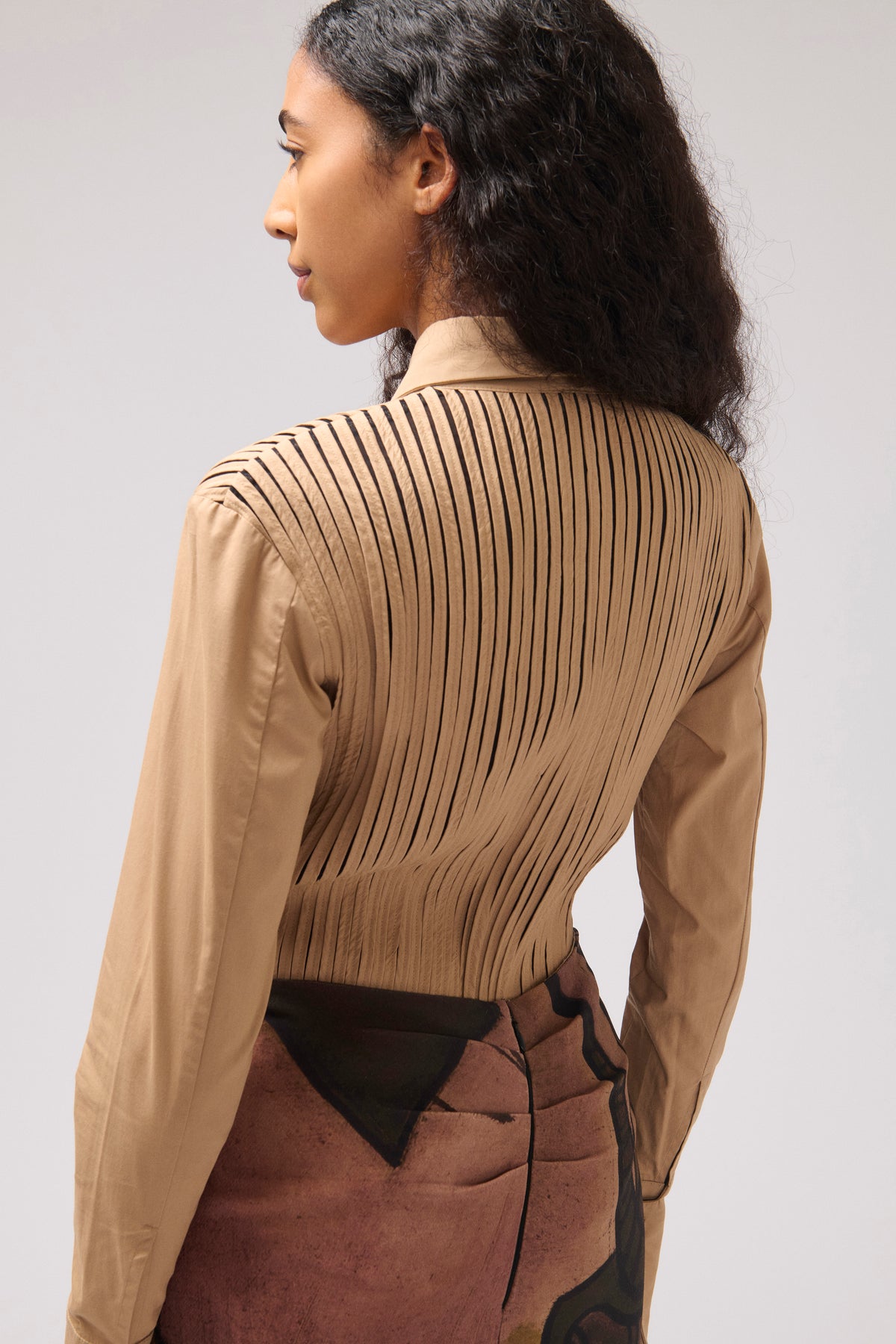 A model shown from the back wearing a beige long-sleeve blouse with vertical pleats across the back and a brown skirt, set against a plain light gray studio background.