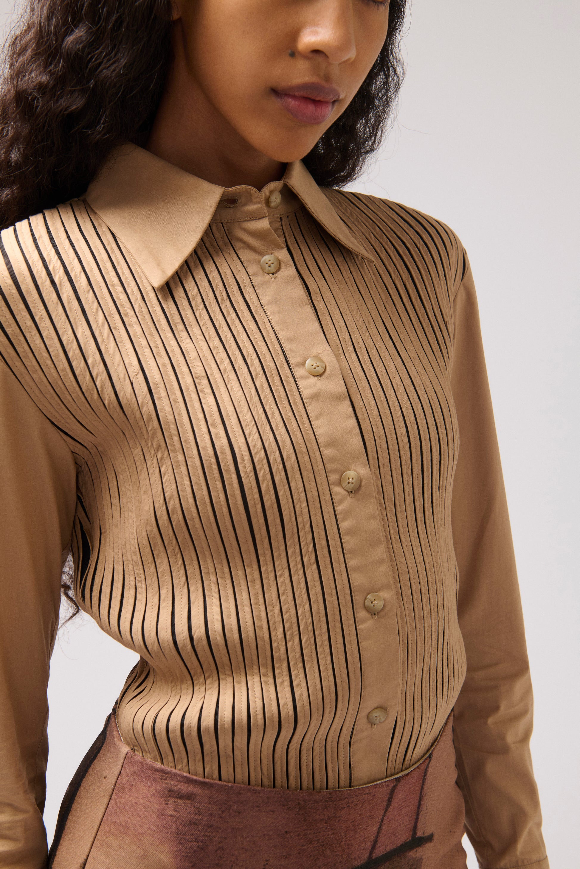 A close-up of a model wearing a tan button-up shirt with vertical black pinstripes and a pointed collar, paired with a brown skirt against a light gray background.