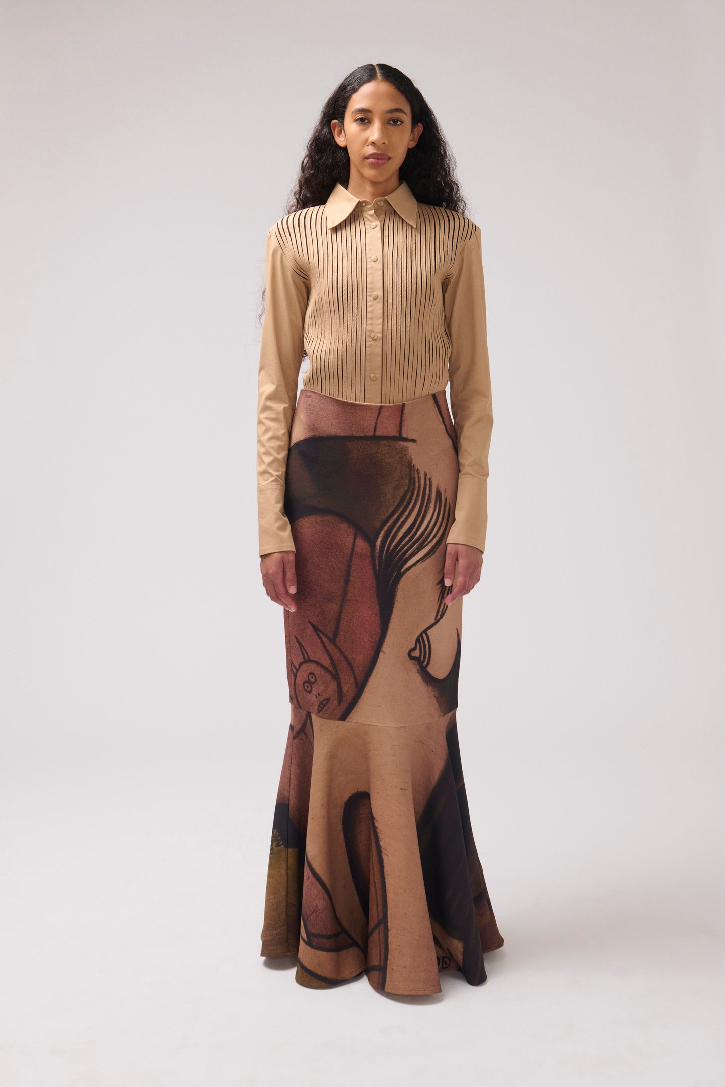A model stands against a light gray studio background wearing a long-sleeve button-up shirt with vertical pleats and a matching long skirt featuring an abstract painterly  print in brown, tan, and black.