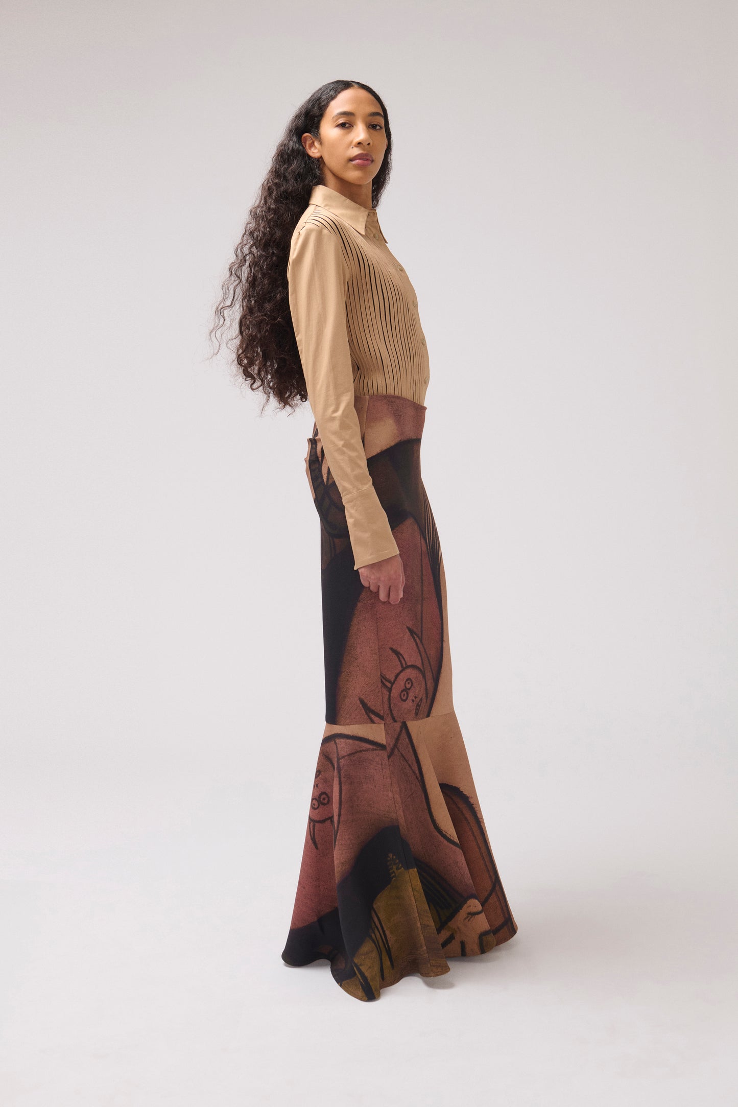A model is shown in profile wearing a beige long-sleeve ribbed crop top and a high-waisted maxi skirt featuring a brown and black abstract graphic print, set against a plain light gray studio background.