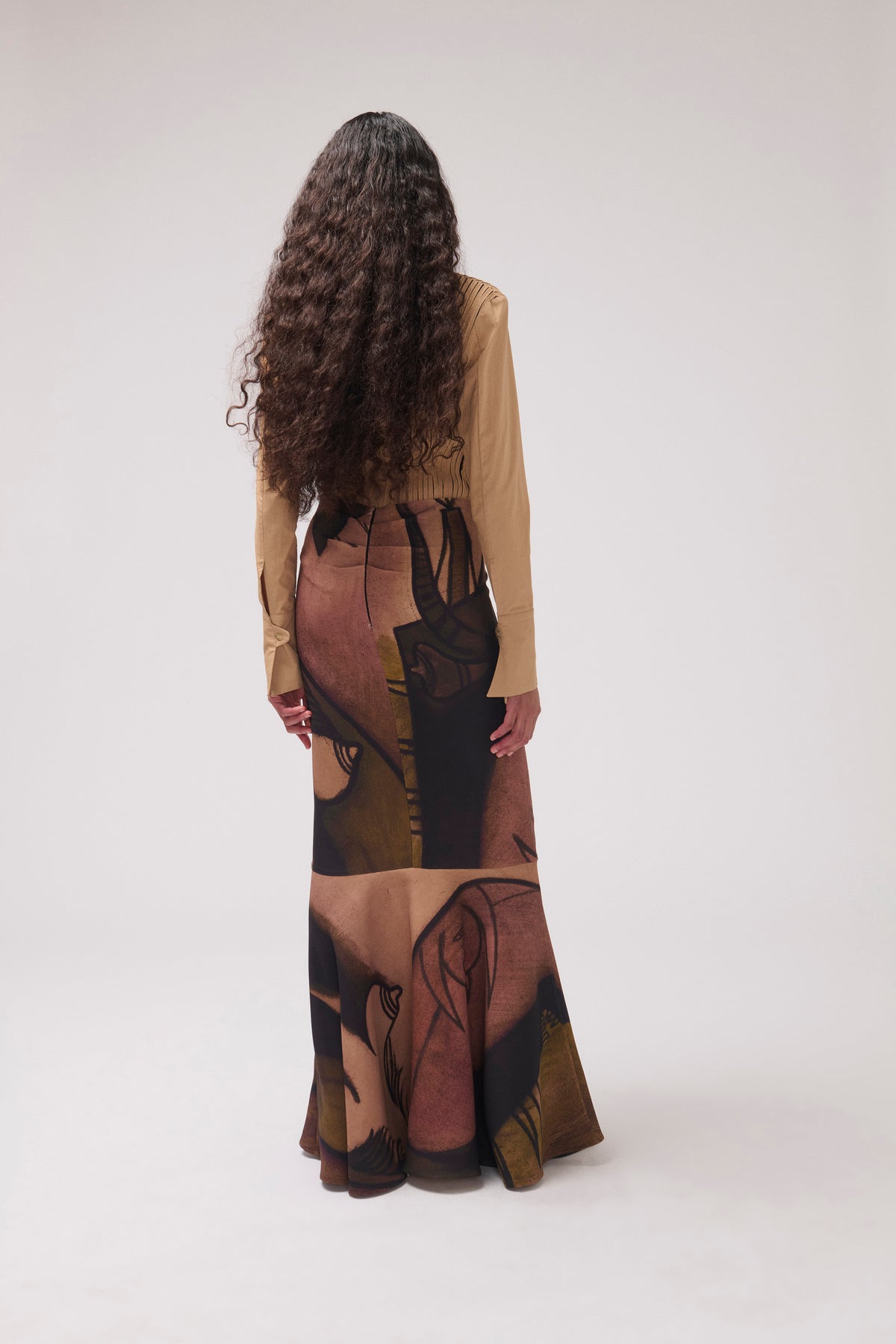 A woman with long, curly dark brown hair is shown from behind, wearing a beige long-sleeve top and a high-waisted maxi skirt with an abstract art print in brown, beige, and black. She is photographed against a seamless, light gray studio background.
