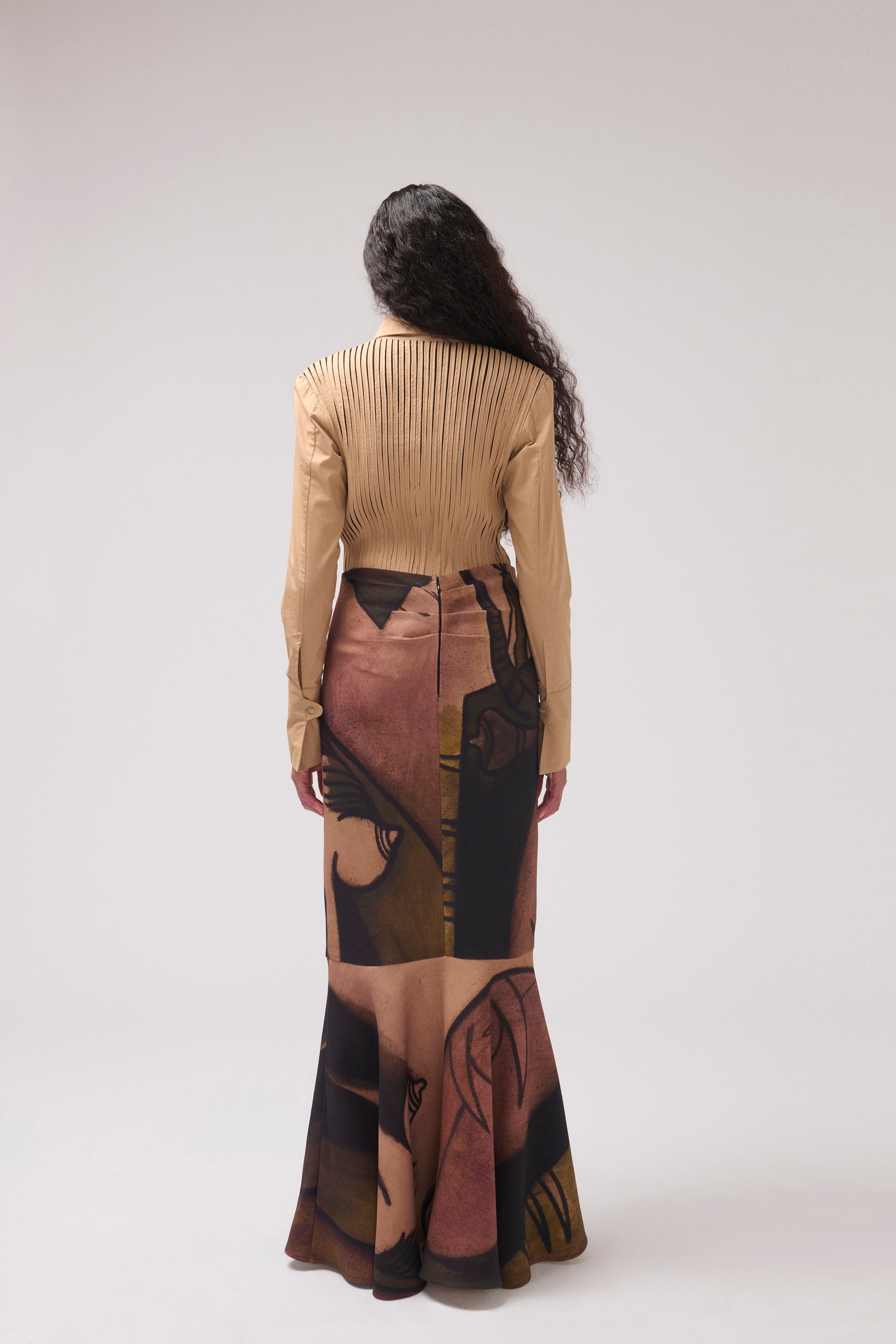 A model viewed from behind wearing a beige long-sleeve pleated top and a long skirt with a large abstract portrait print in beige, brown, and black, against a light gray studio background.