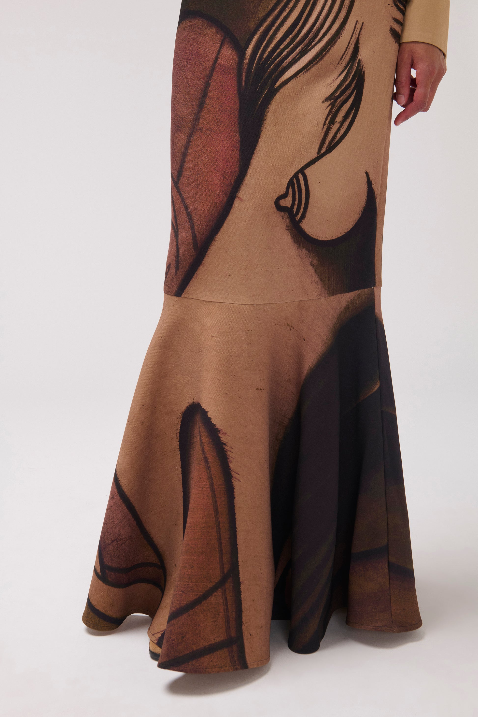 A cropped image of a model standing against a light gray studio background wearing a long-sleeve button-up shirt with vertical pleats and a matching long skirt featuring an abstract painterly  print in brown, tan, and black.