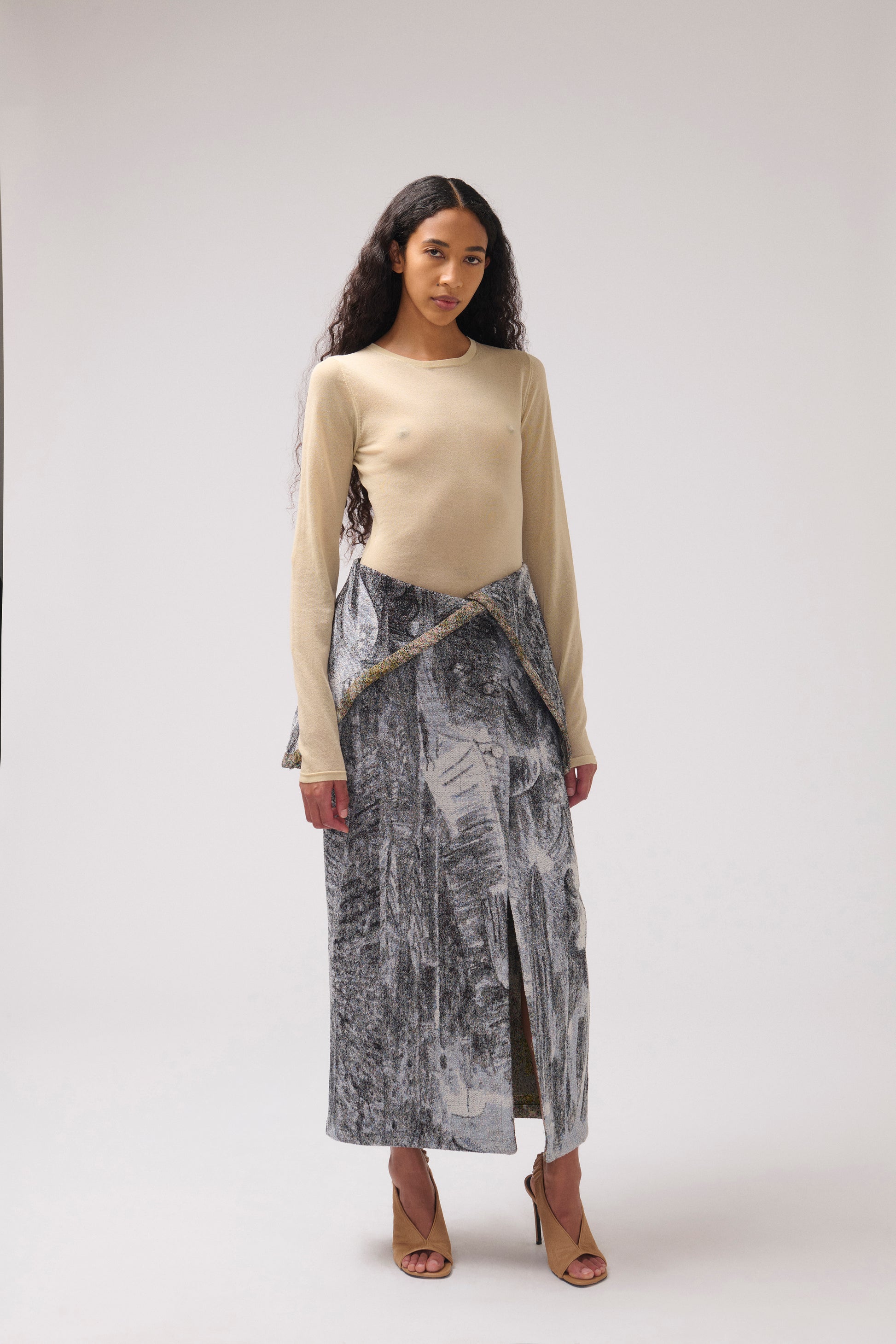 A model stands against a light gray studio background wearing a  long draped skirt with a multi-color print. 