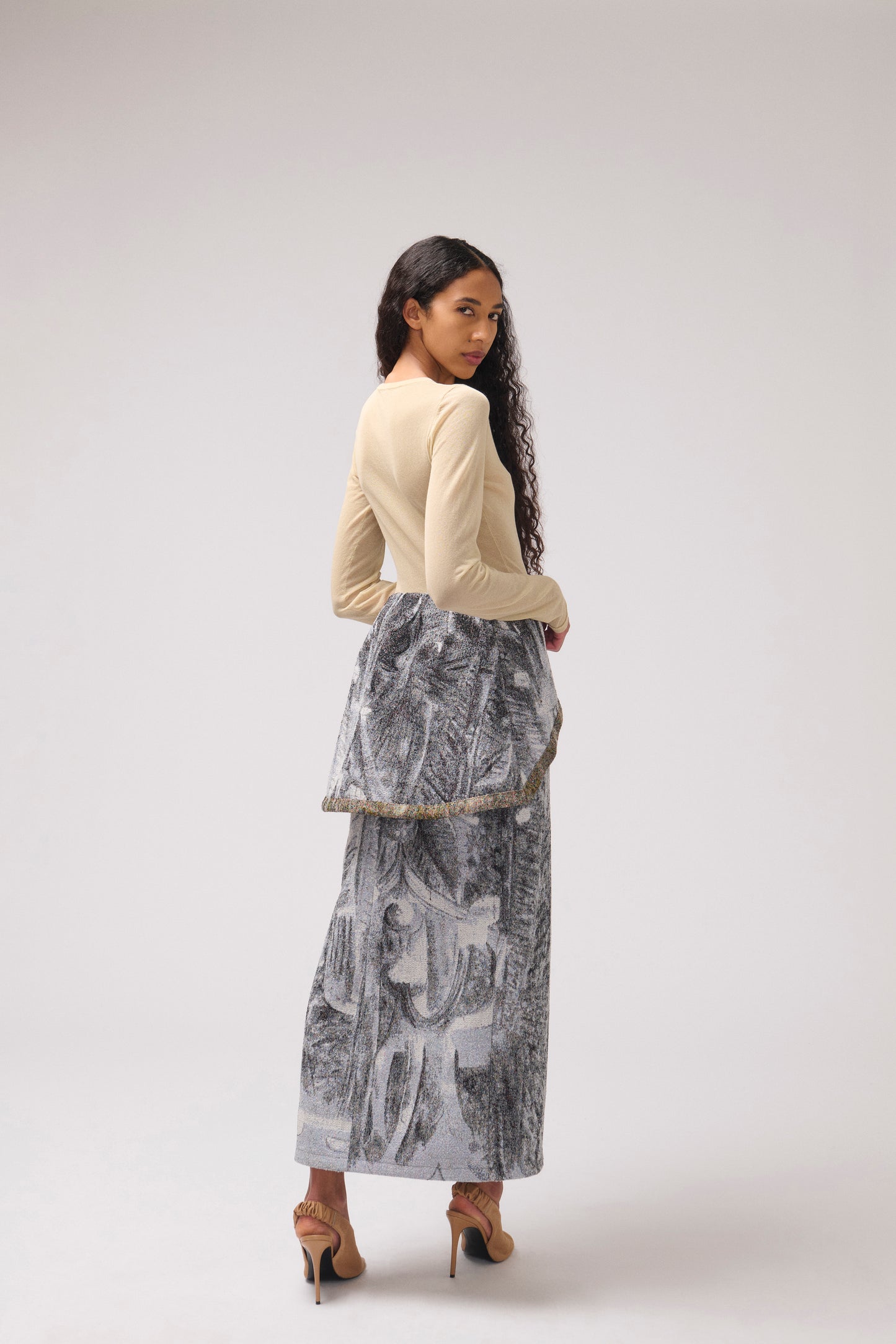 A model is shown from behind wearing a  long draped skirt with a multi-color print. The model is standing against a light gray studio background.